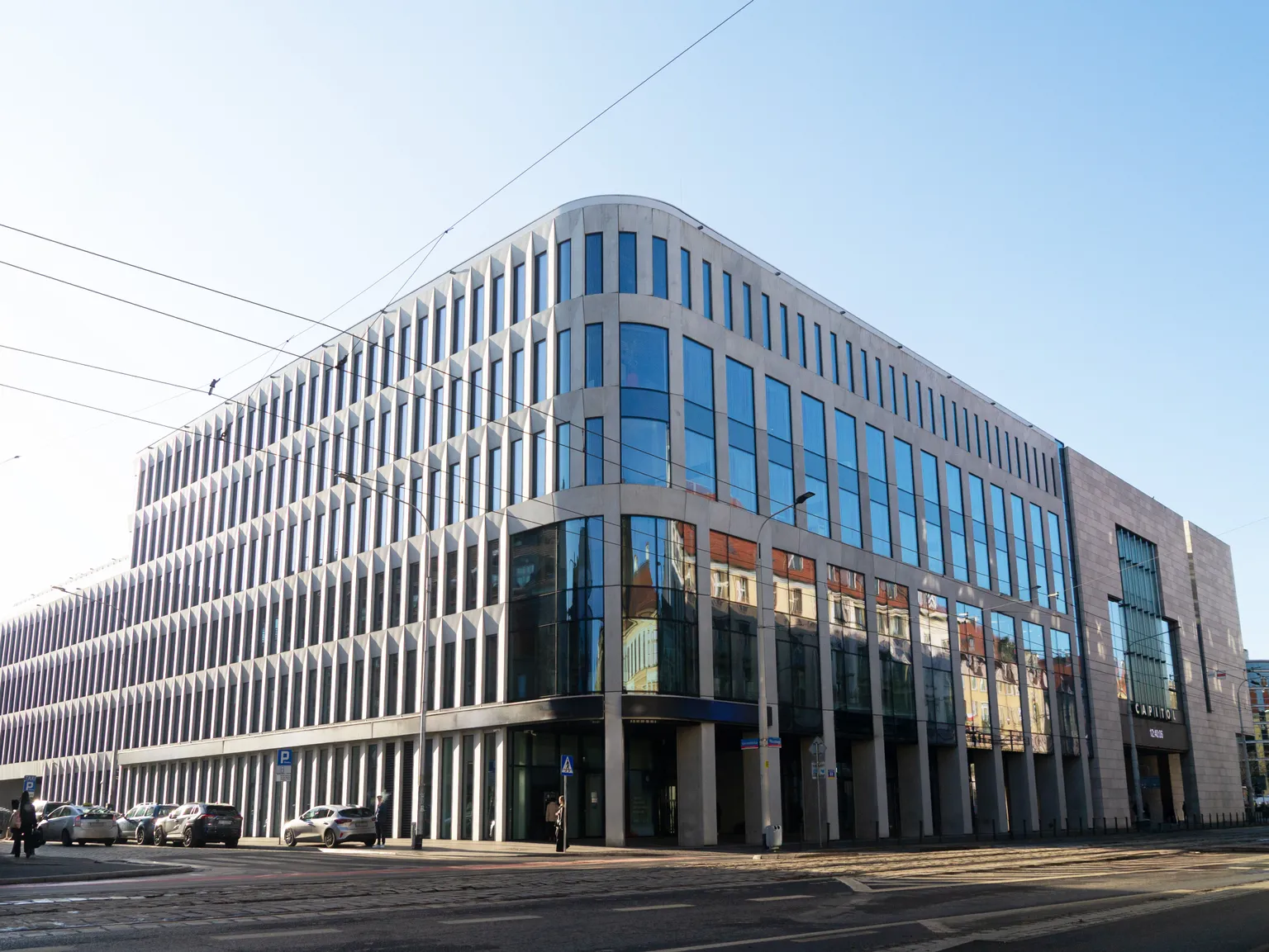 Pratia Wrocław Oncology -Poland_ Modern building with large reflective windows, rows of vertical columns, and parked cars along the street under a clear blue sky.