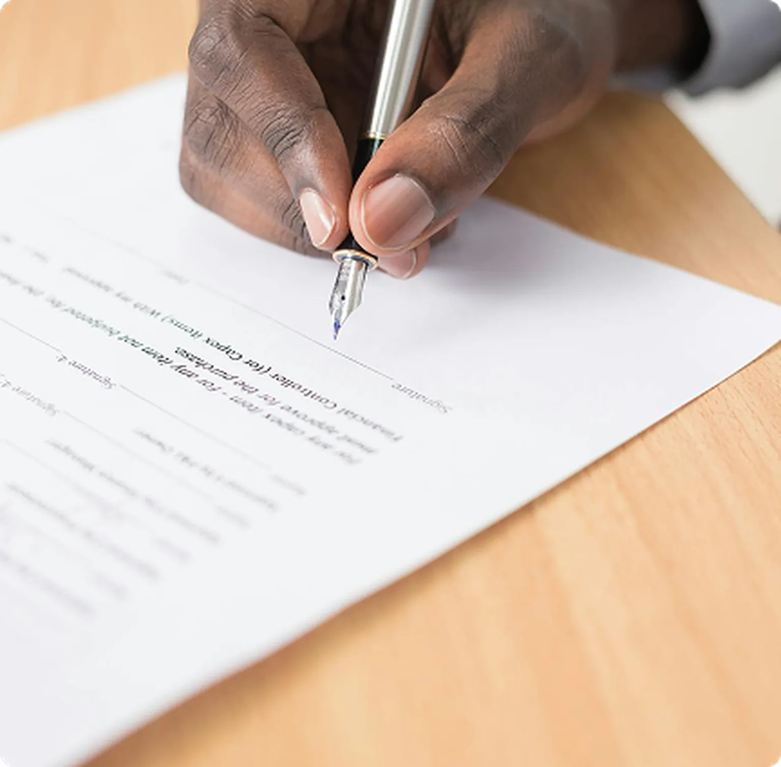 A hand holding a pen poised to sign or write on a piece of printed paper on a wooden table.