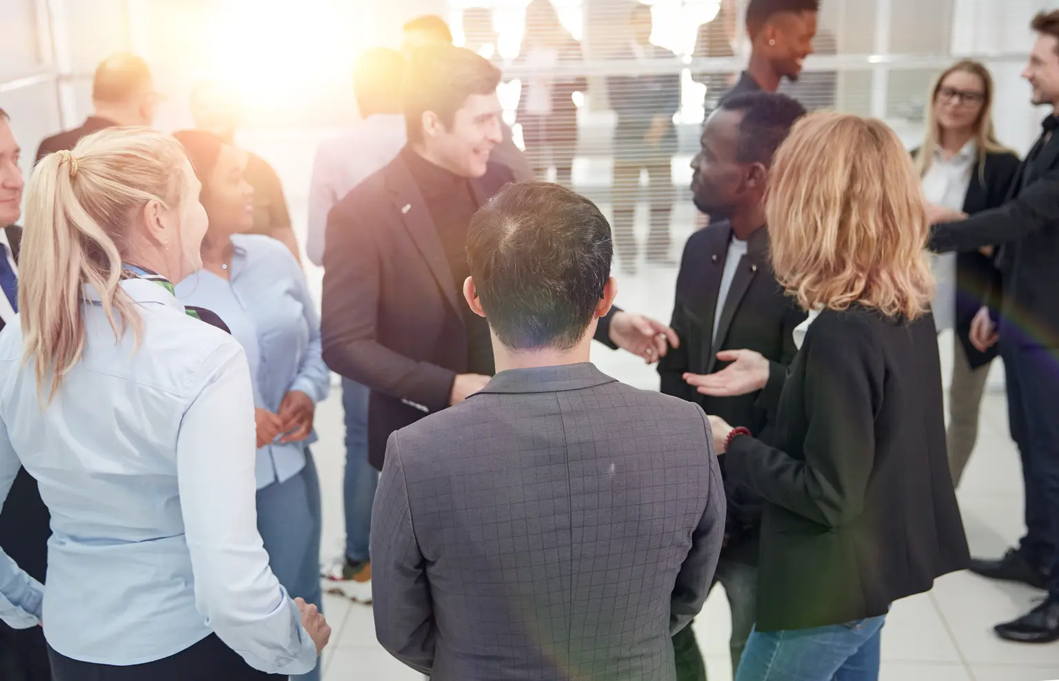 A diverse group of people in business attire are engaged in lively conversation in a bright, modern office setting.