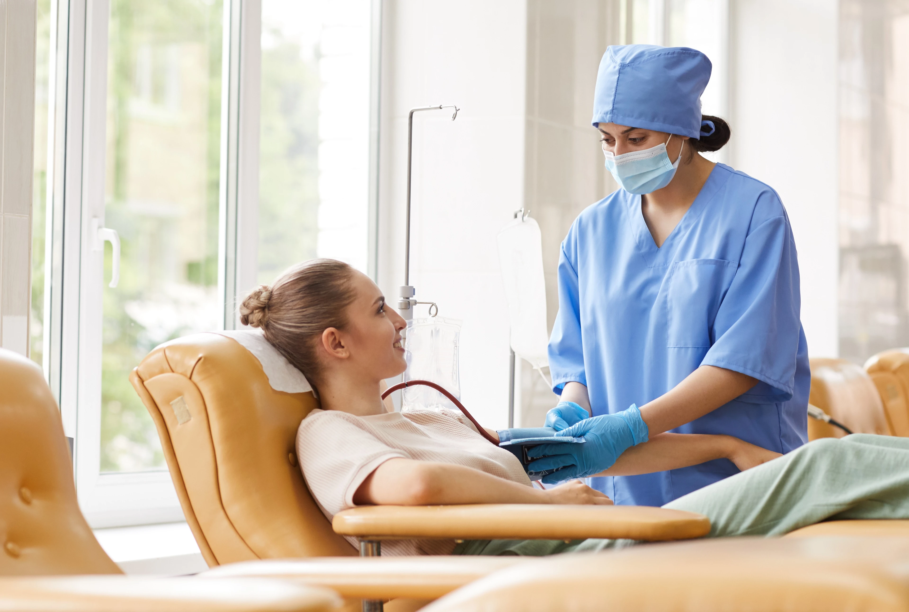 A healthcare worker in blue scrubs and mask assists a woman donating blood in a clinic, using a device to monitor her vitals.