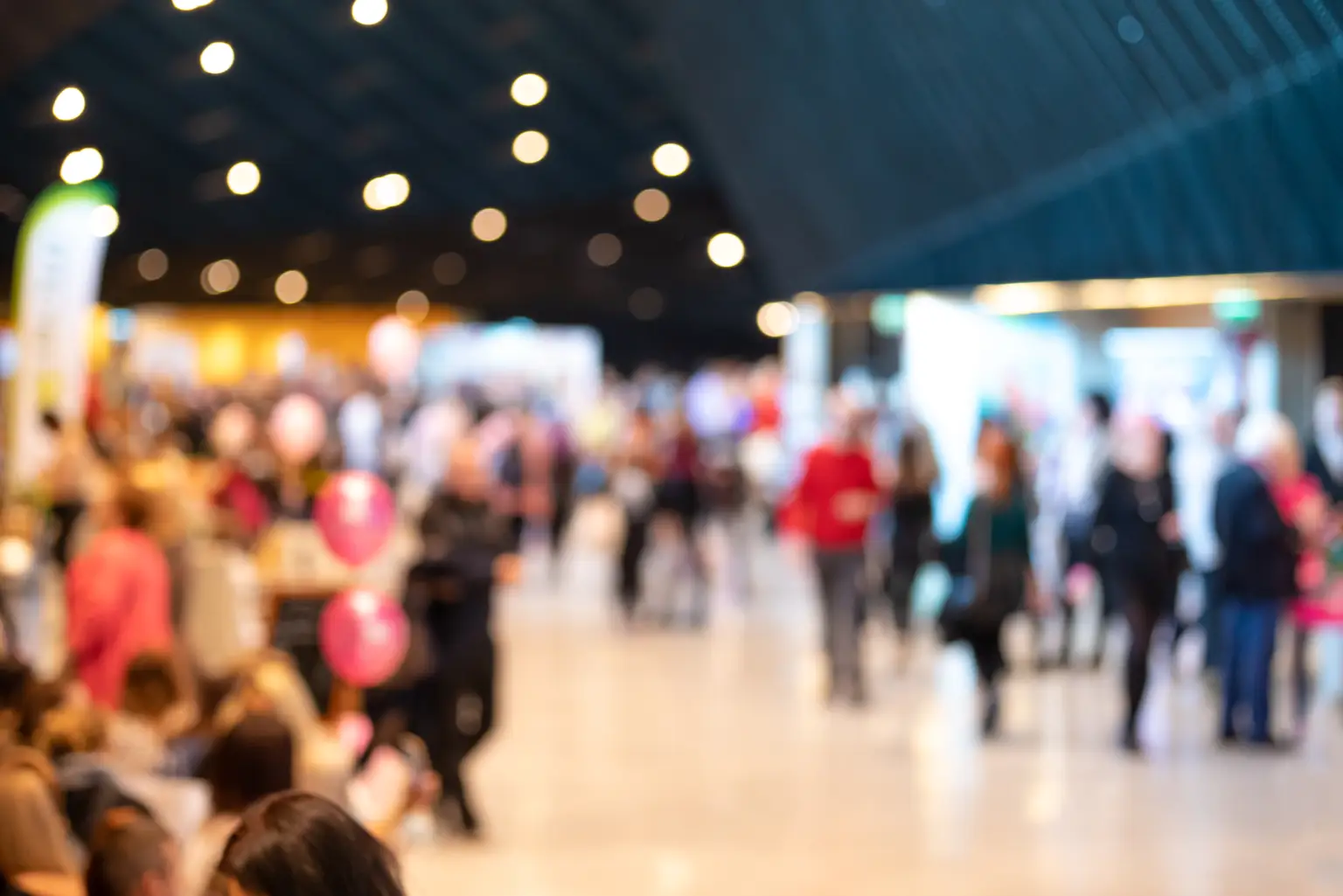 Blurred image of a busy indoor event with people walking around, bright overhead lights, and colorful stands.