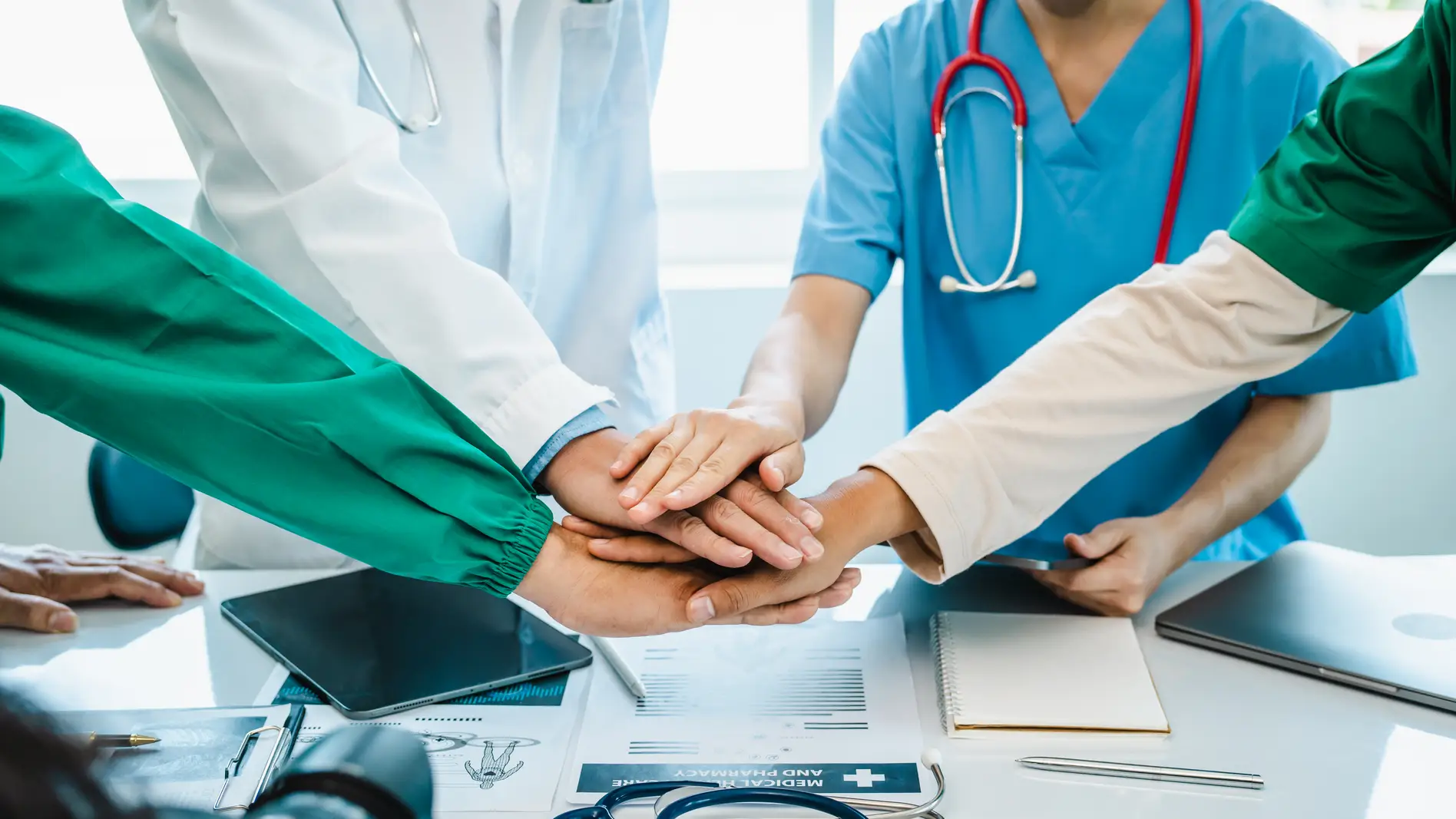 Healthcare professionals in uniforms stack hands in teamwork over a table with medical documents and equipment.