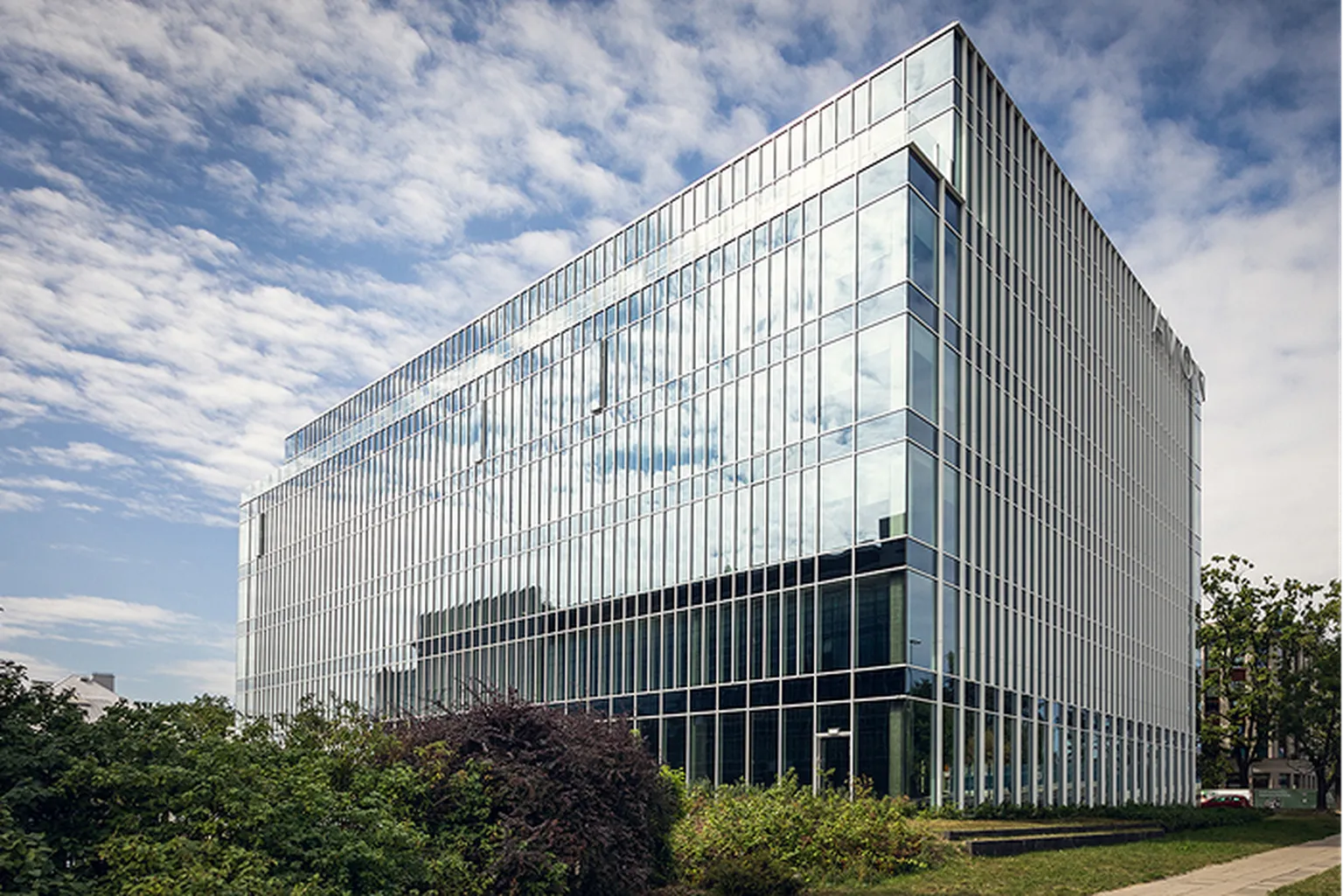 Modern glass office building with reflective windows, surrounded by greenery, under a partly cloudy sky.