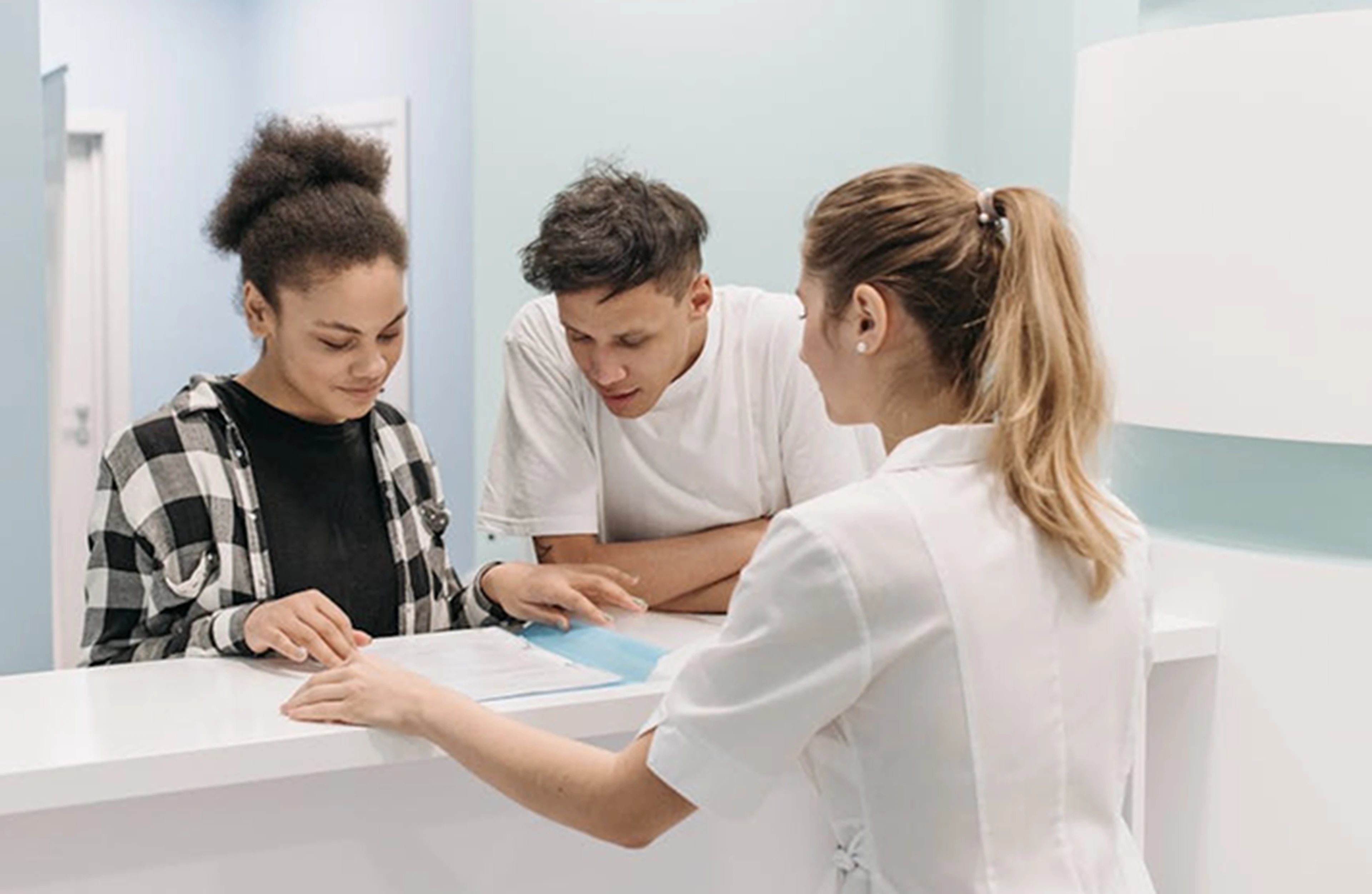 Two people sign documents at a reception desk, assisted by a female staff member in a white uniform, in a bright, modern office space.