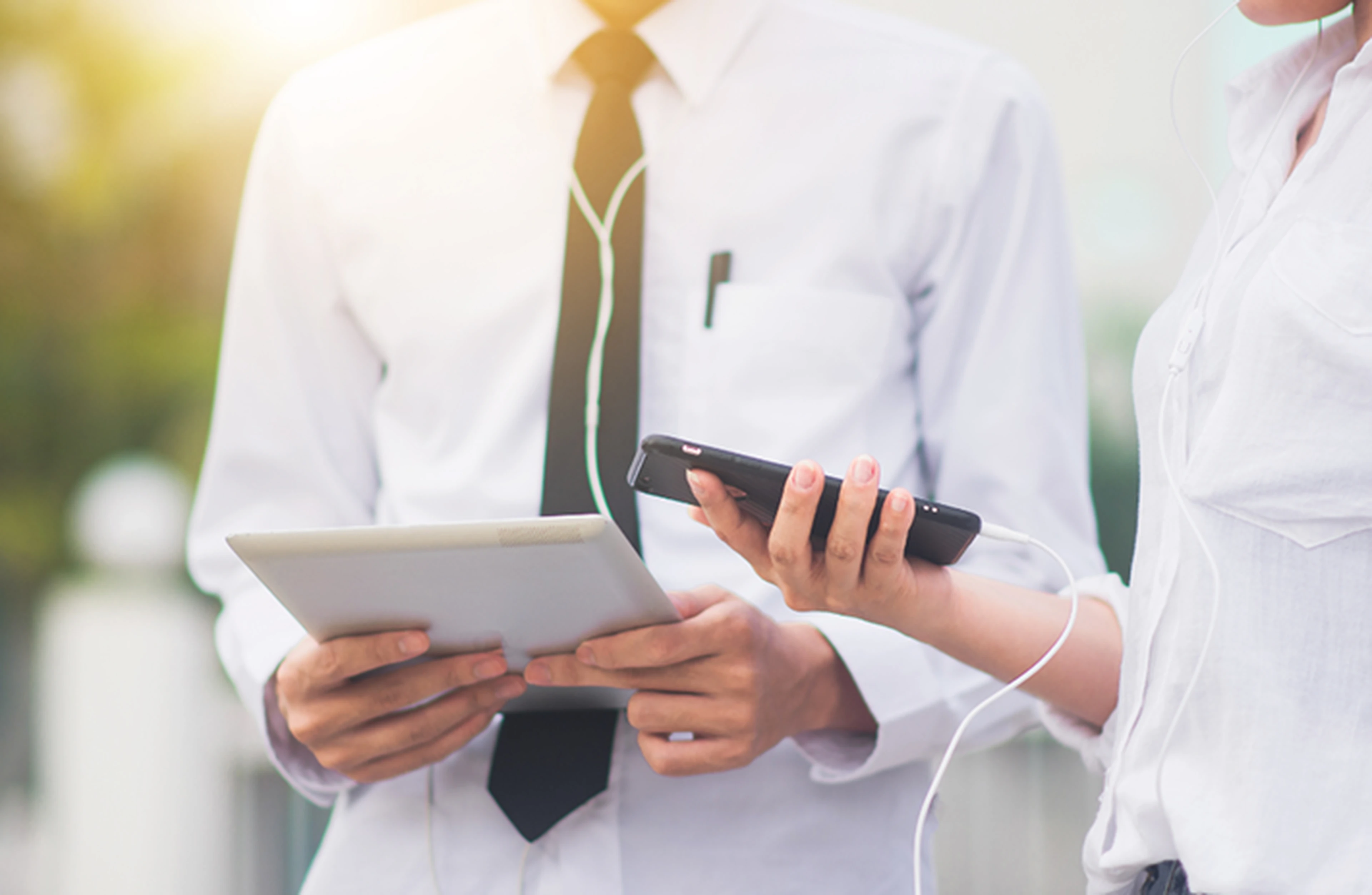 Two people in white shirts sharing earphones, one holding a tablet and the other a smartphone. Bright, outdoor setting.