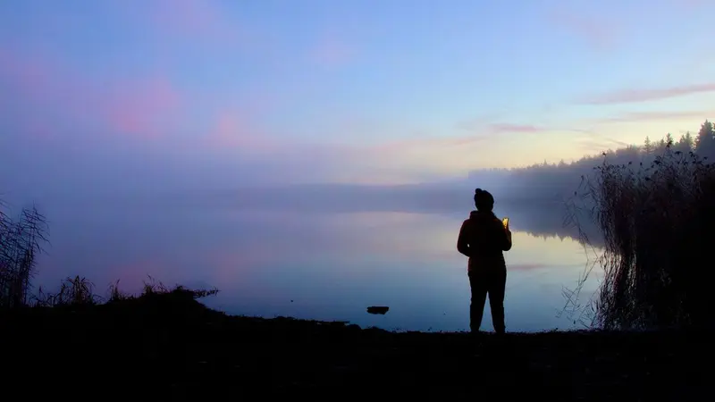 Lone person standing by calm water at dusk