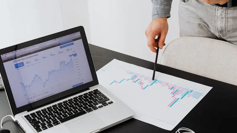 Laptop showing a chart while a hand points at printed graphs
