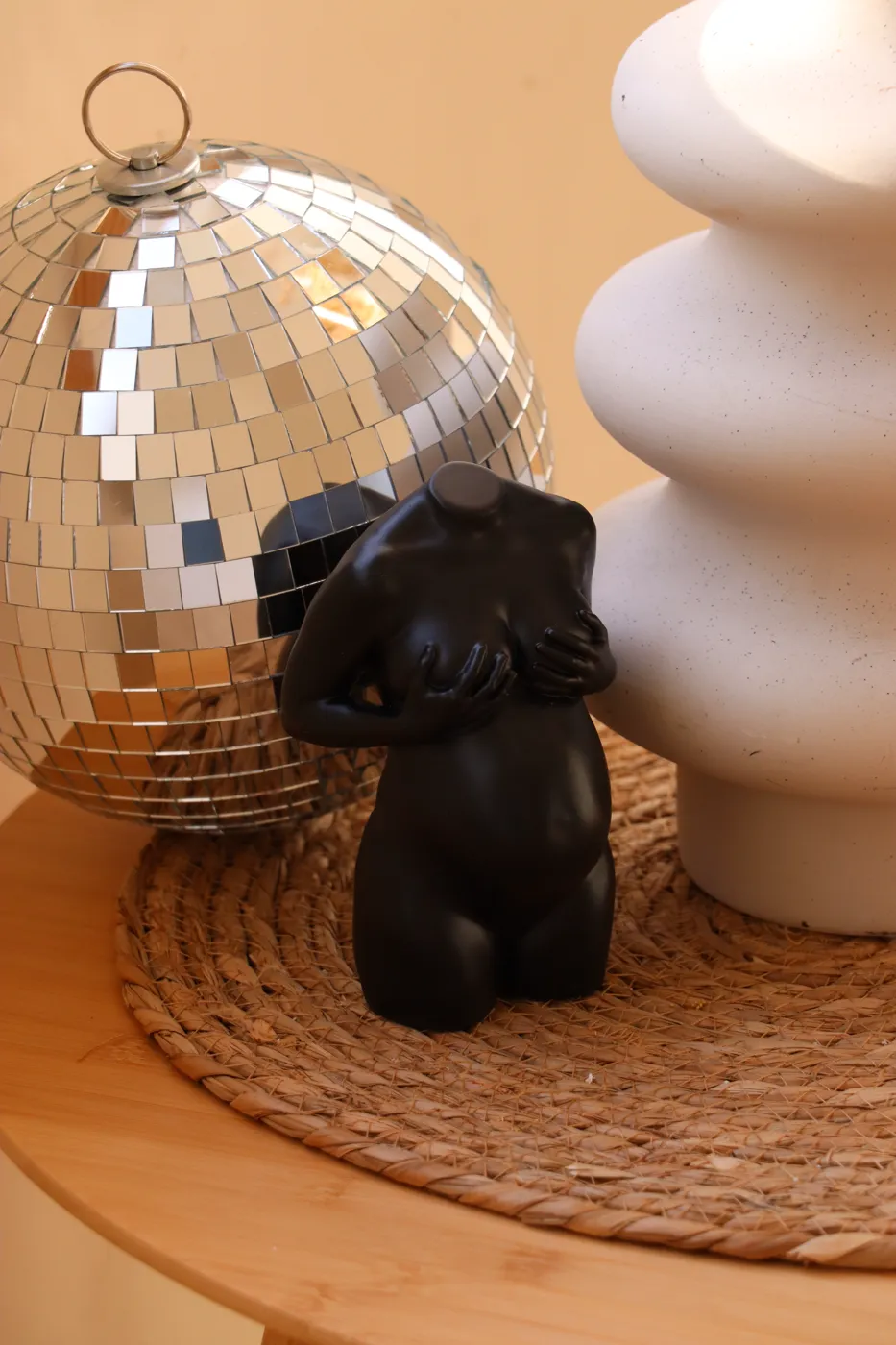 Headless black statue of a pregnant woman, with a disco ball and white ceramic piece on a woven mat on a wooden table.