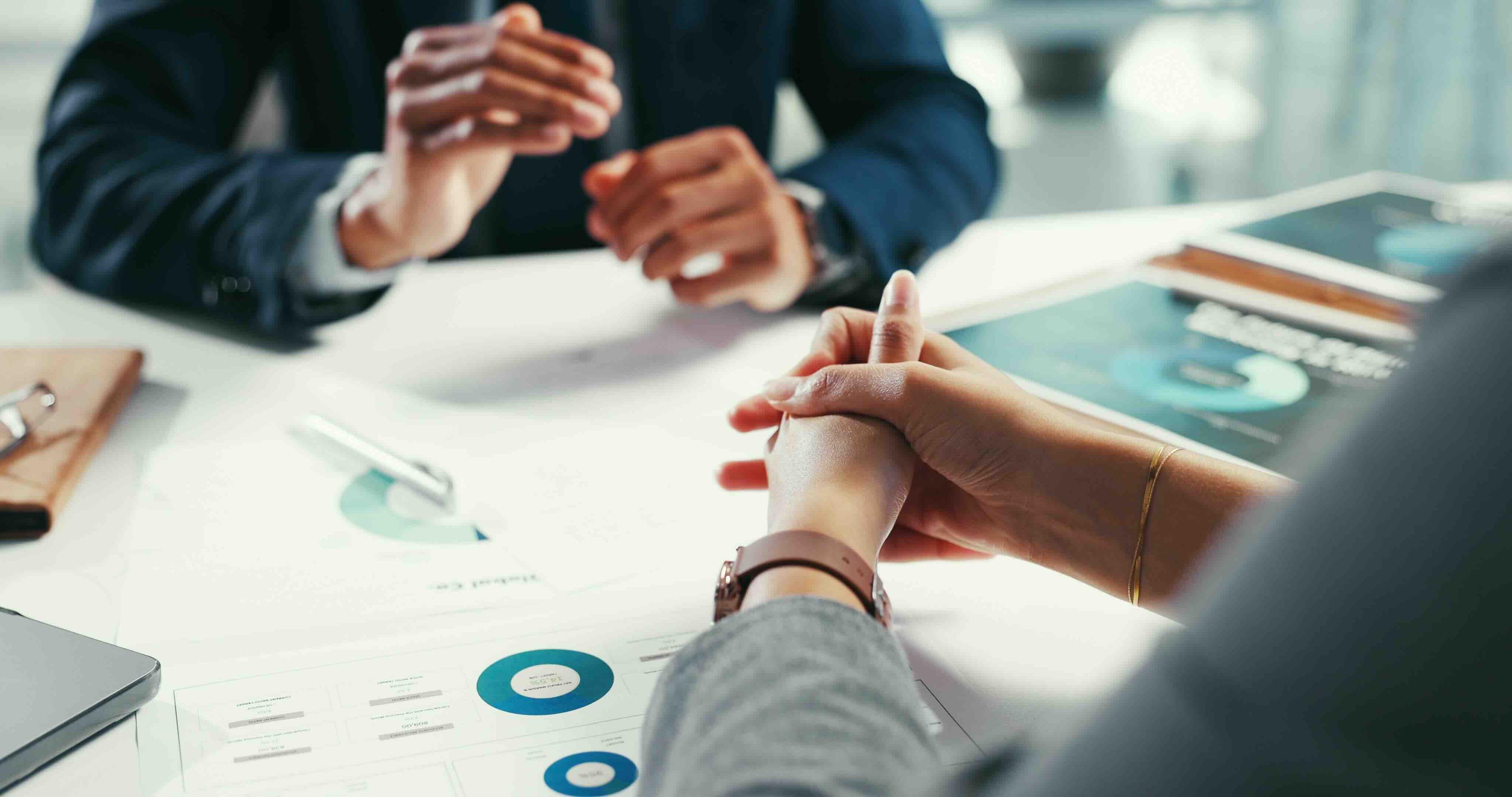 Two people in business attire discussing over documents and charts on a table. Hands are focused, suggesting a professional meeting.