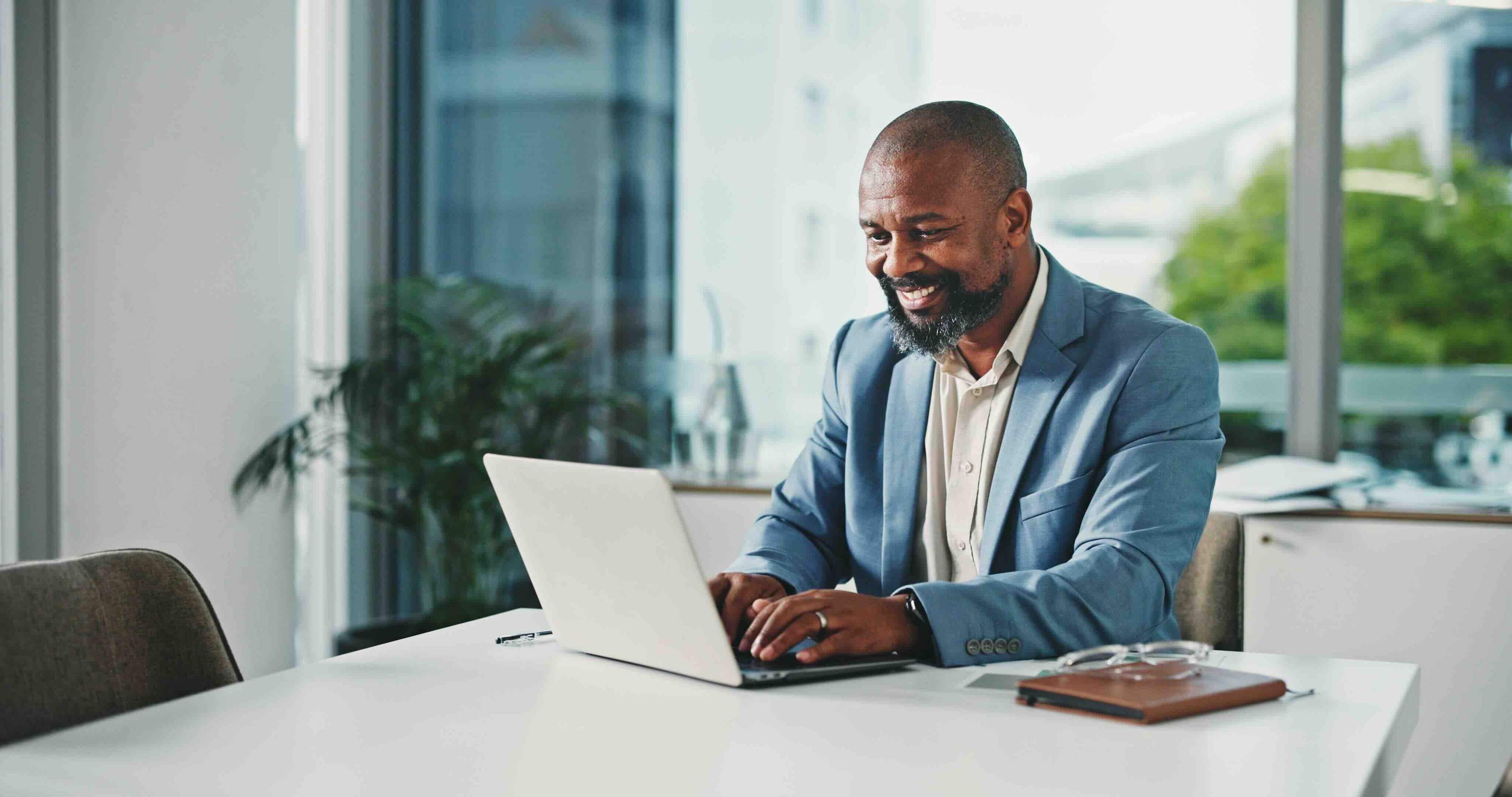 Man in a blue blazer smiling at a laptop in a modern office, with large windows and greenery outside.