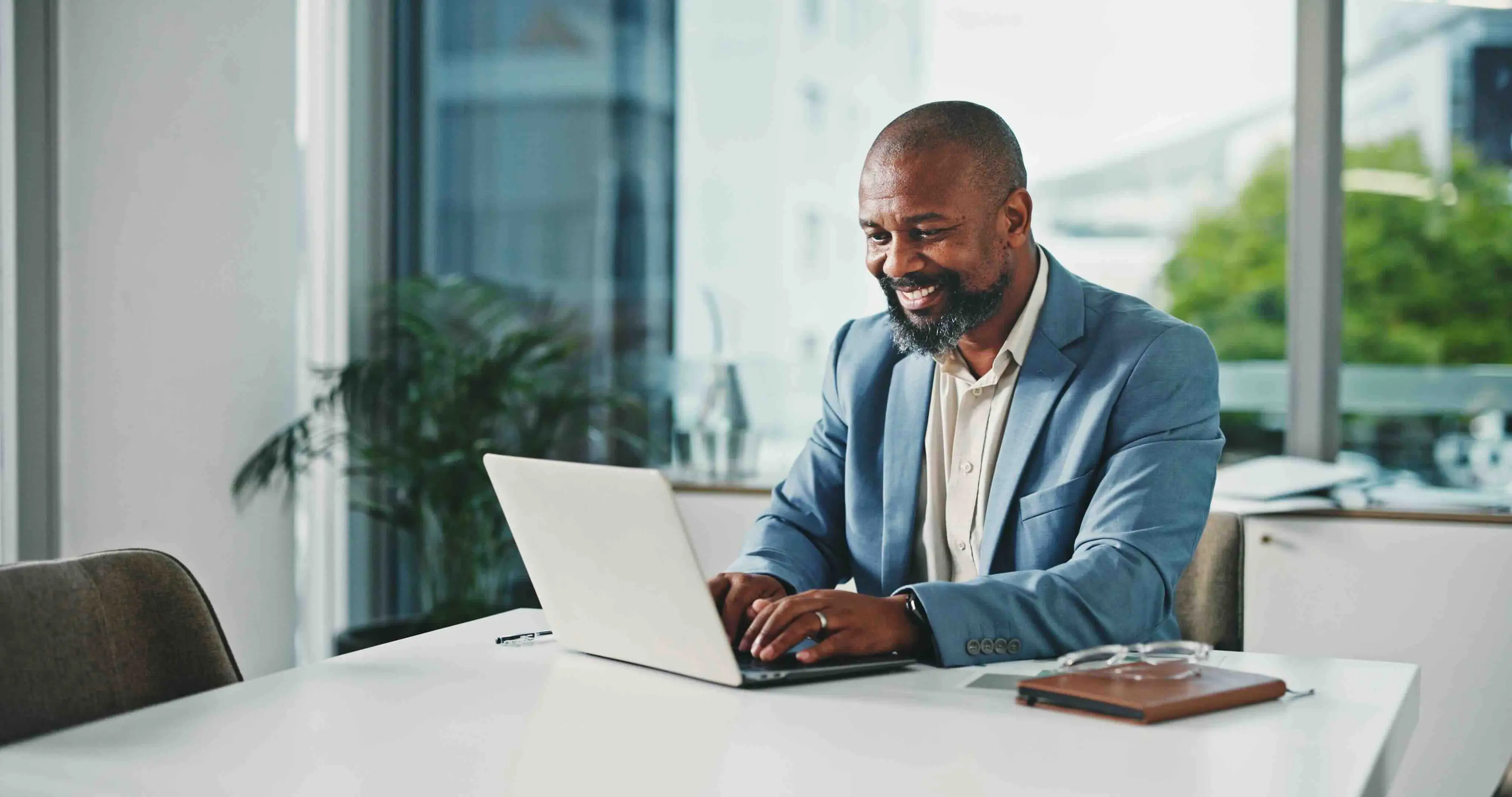 Man in a blue blazer smiling at a laptop in a modern office, with large windows and greenery outside.