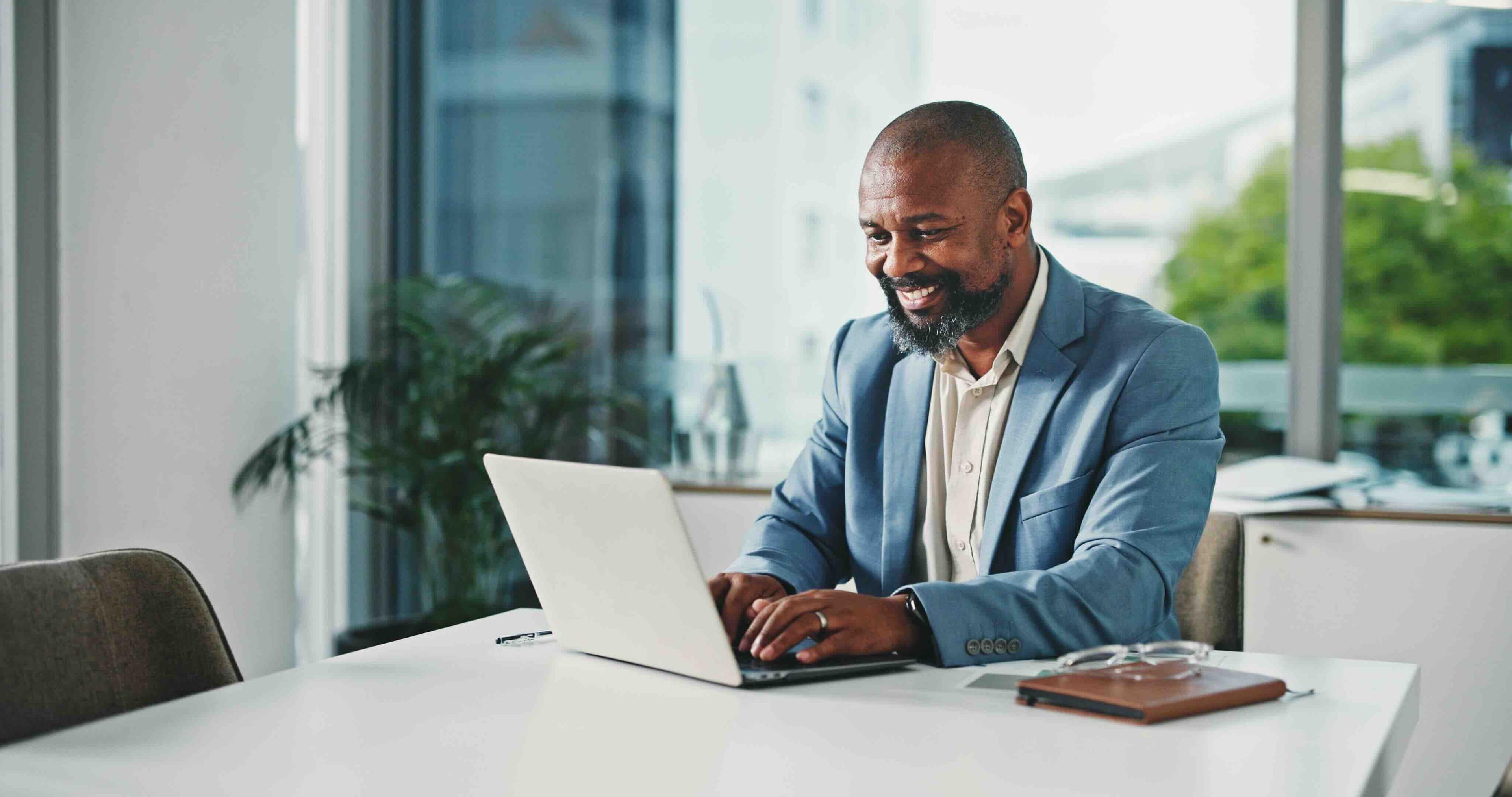 Man in a blue blazer smiling at a laptop in a modern office, with large windows and greenery outside.