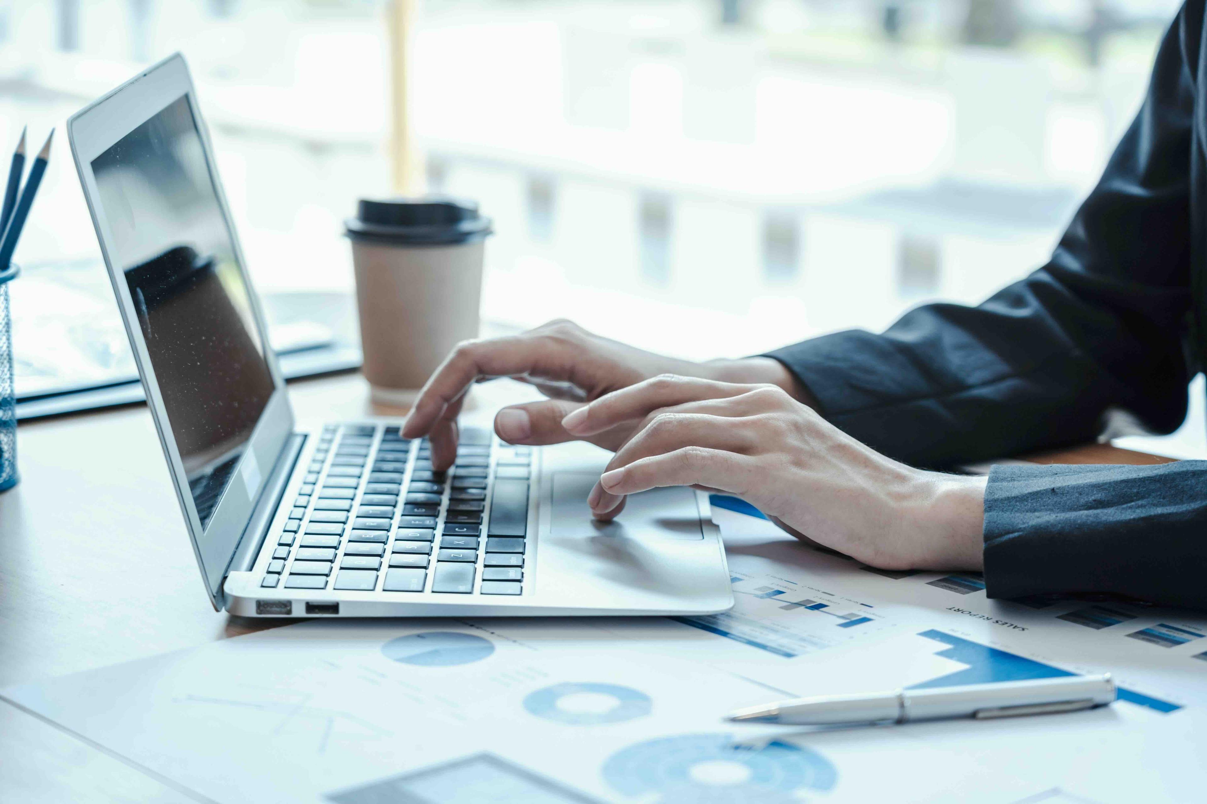 Person typing on a laptop at a desk with charts, coffee cup, and pen, indicating a work or study setting.
