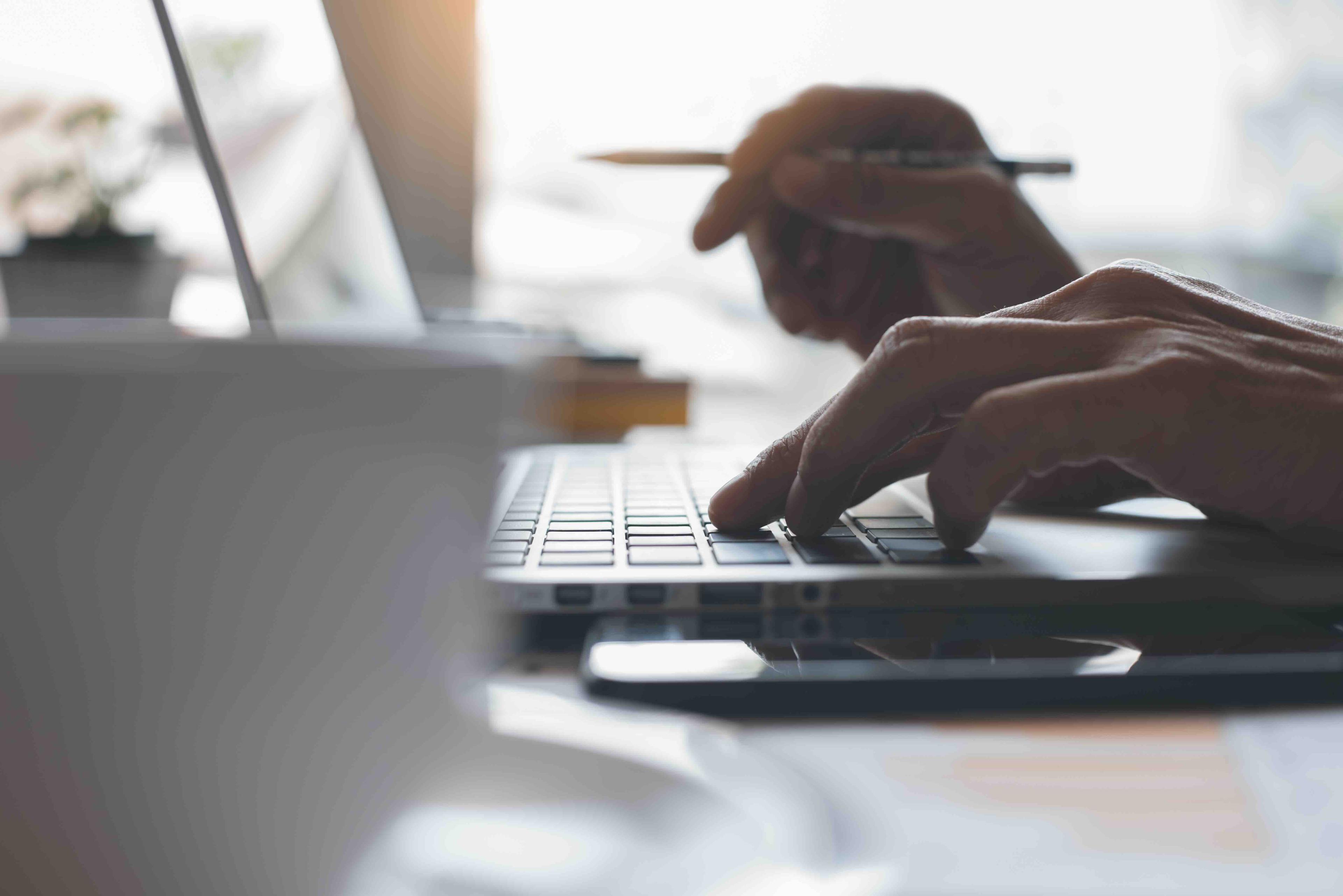 Person typing on a laptop while holding a pen, with a blurred cup and smartphone in the foreground. Sunlight streams through a window.