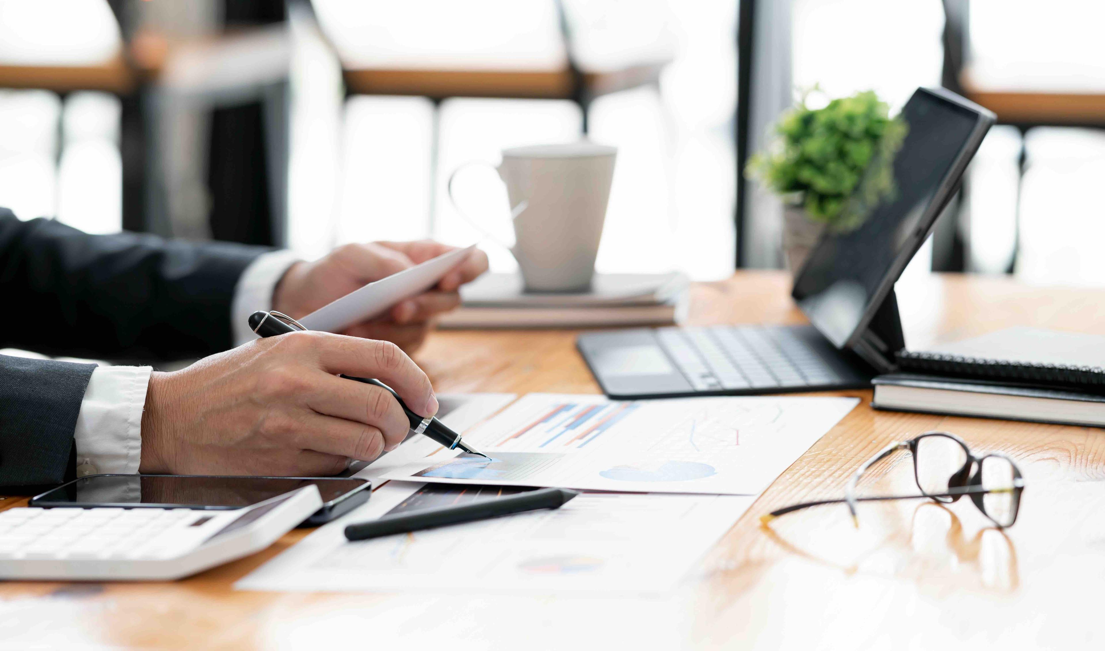 Person reviewing documents with a tablet, coffee, and plant on a wooden desk, accompanied by glasses and calculator.