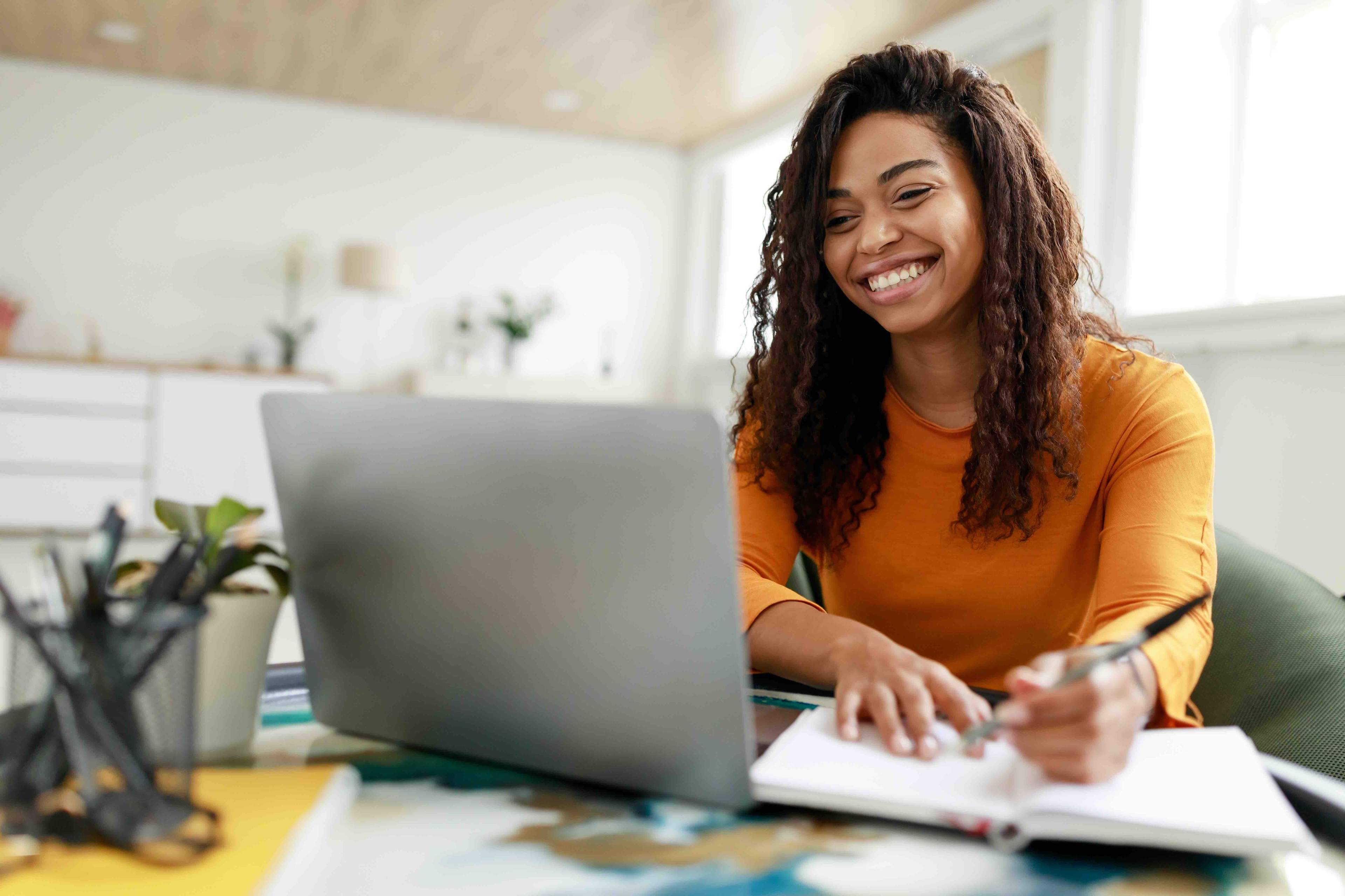 Woman in an orange shirt smiles while working on a laptop, taking notes in a notebook, in a bright home office setting.