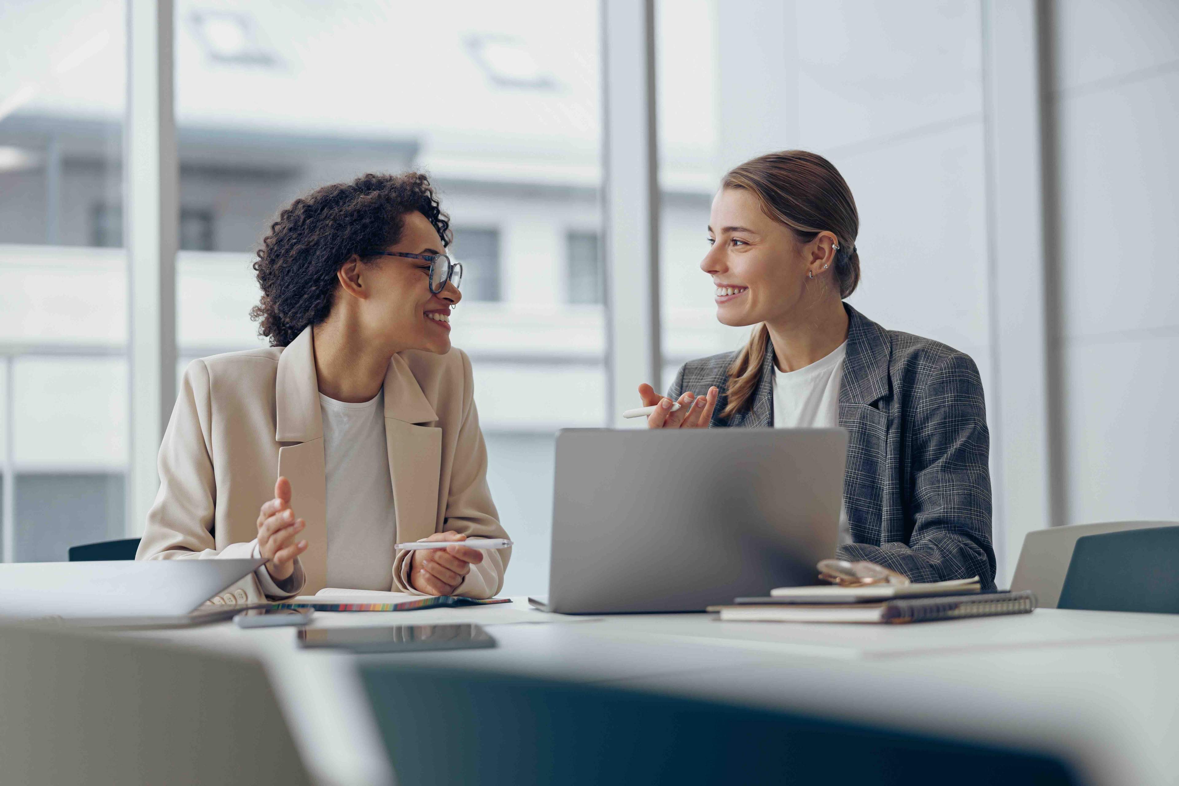 Two professionals sitting at a table, engaged in conversation, with a laptop and notebooks in front of them. Bright office environment.