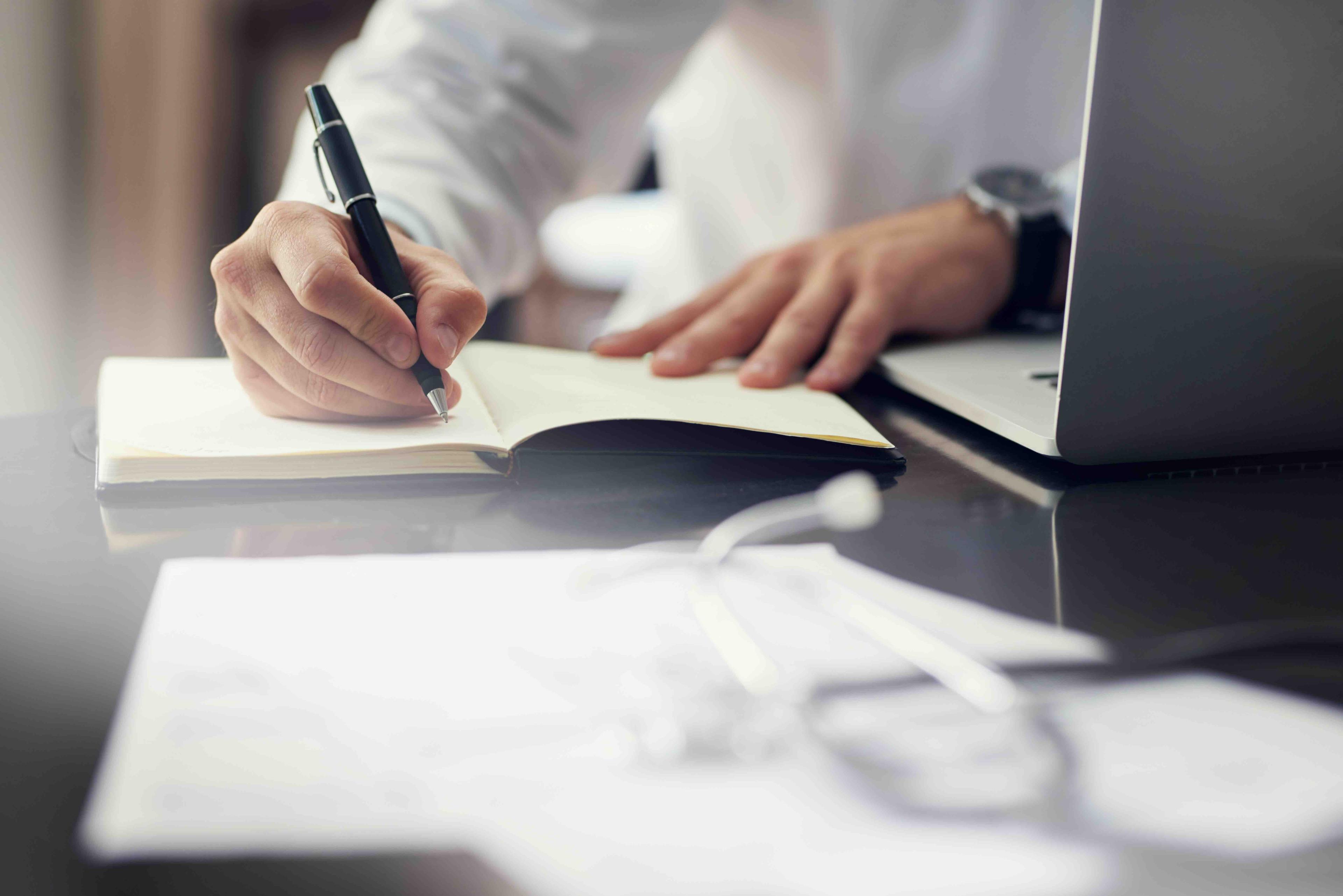 Person writing in a notebook with a pen beside a laptop, on a desk with blurred documents in the foreground.