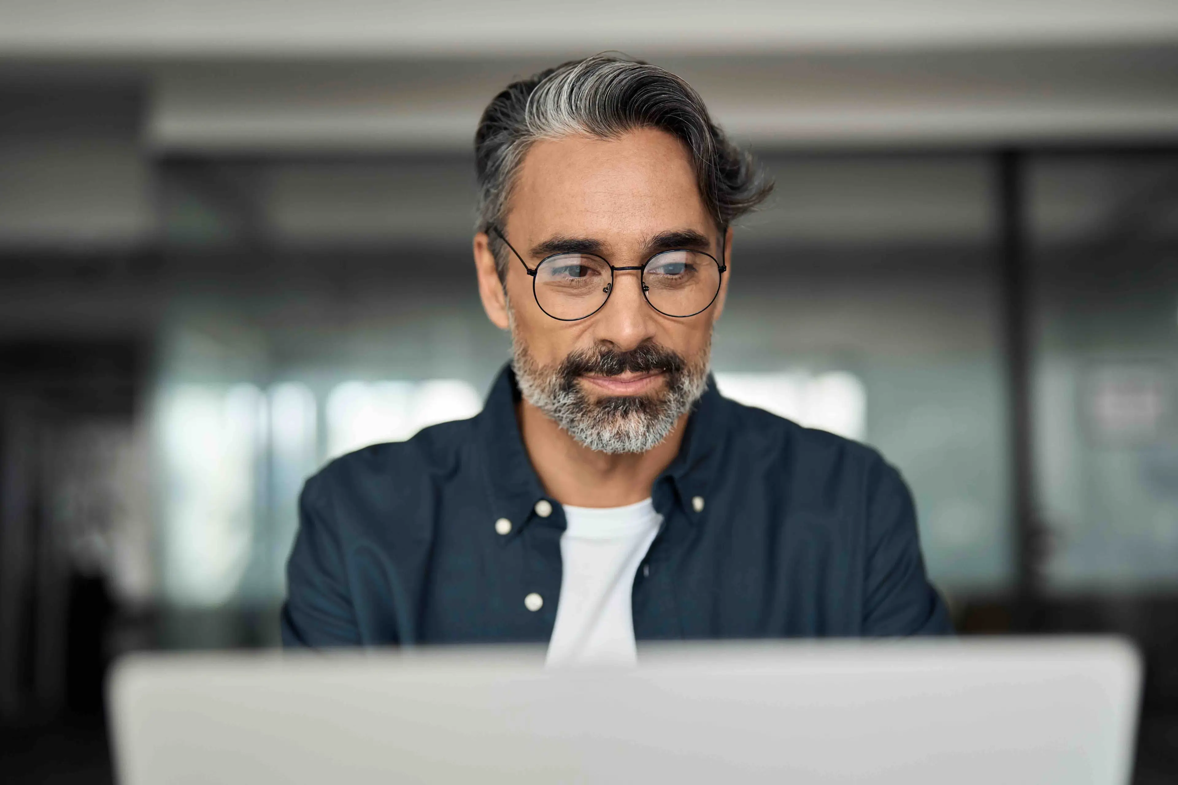 Man with glasses and salt-and-pepper hair using a laptop in a modern, softly-lit office setting, wearing a dark shirt and a white undershirt.