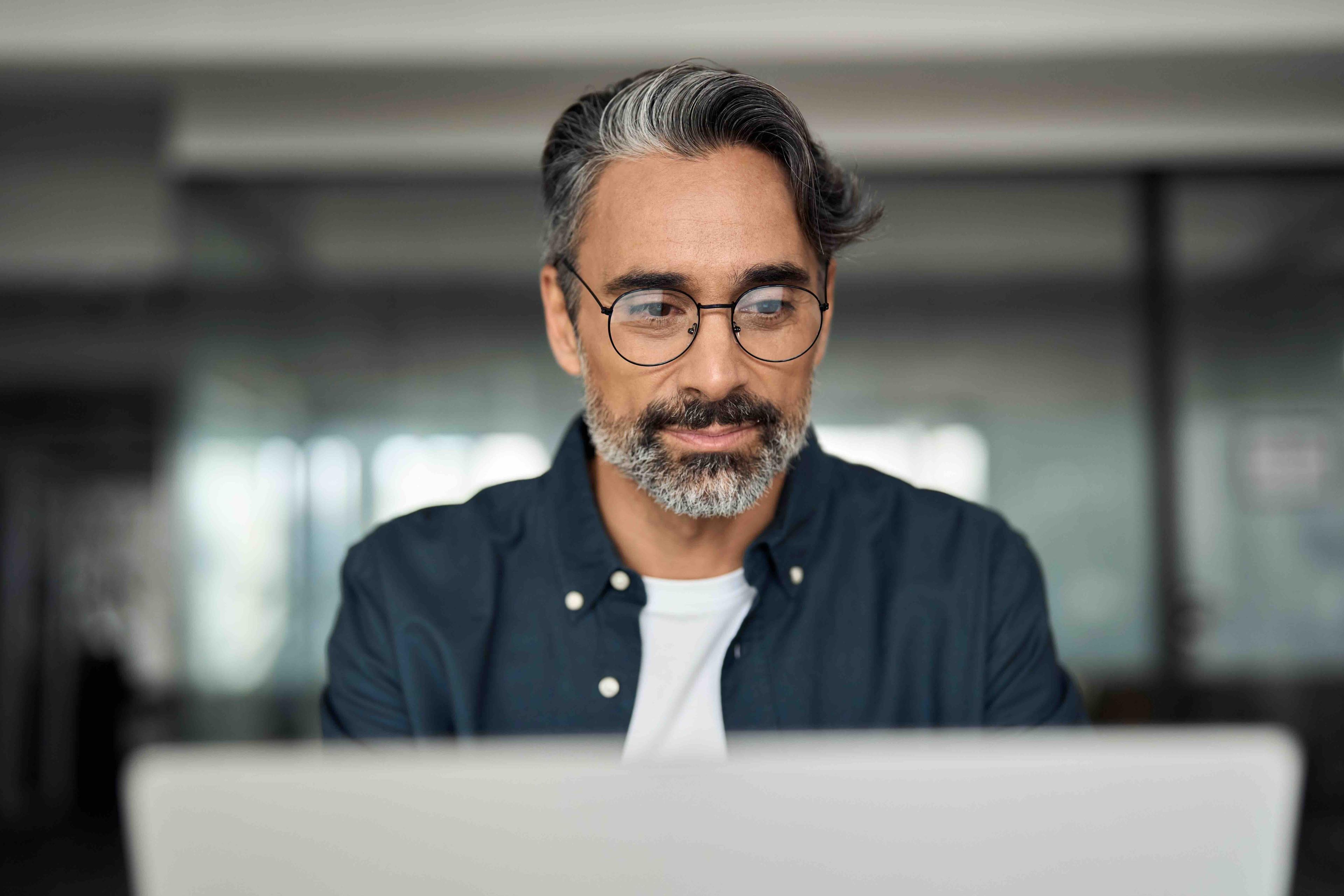 Man with glasses and salt-and-pepper hair using a laptop in a modern, softly-lit office setting, wearing a dark shirt and a white undershirt.