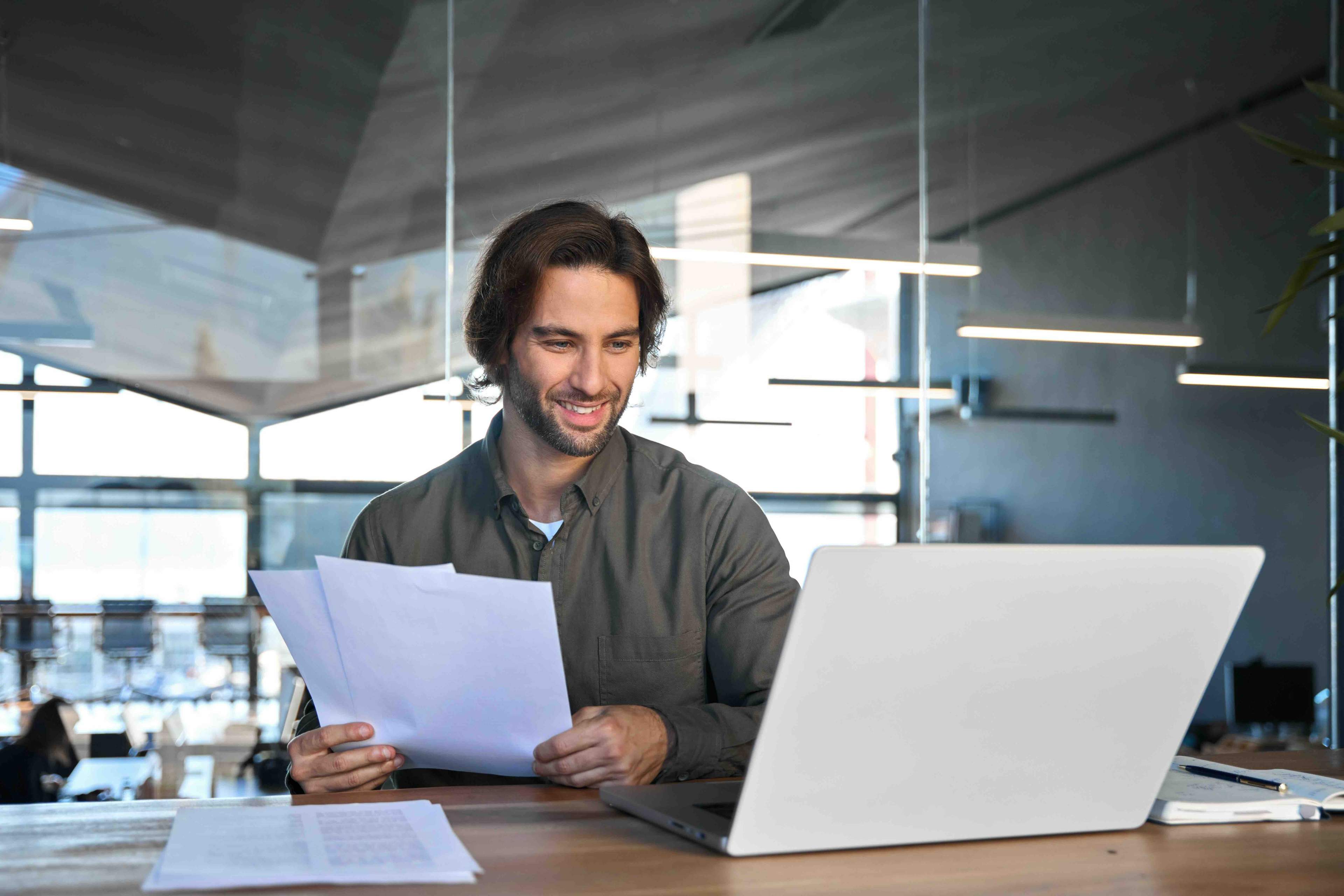 Smiling man holds papers while working on a laptop in a modern office with large windows and bright lighting.