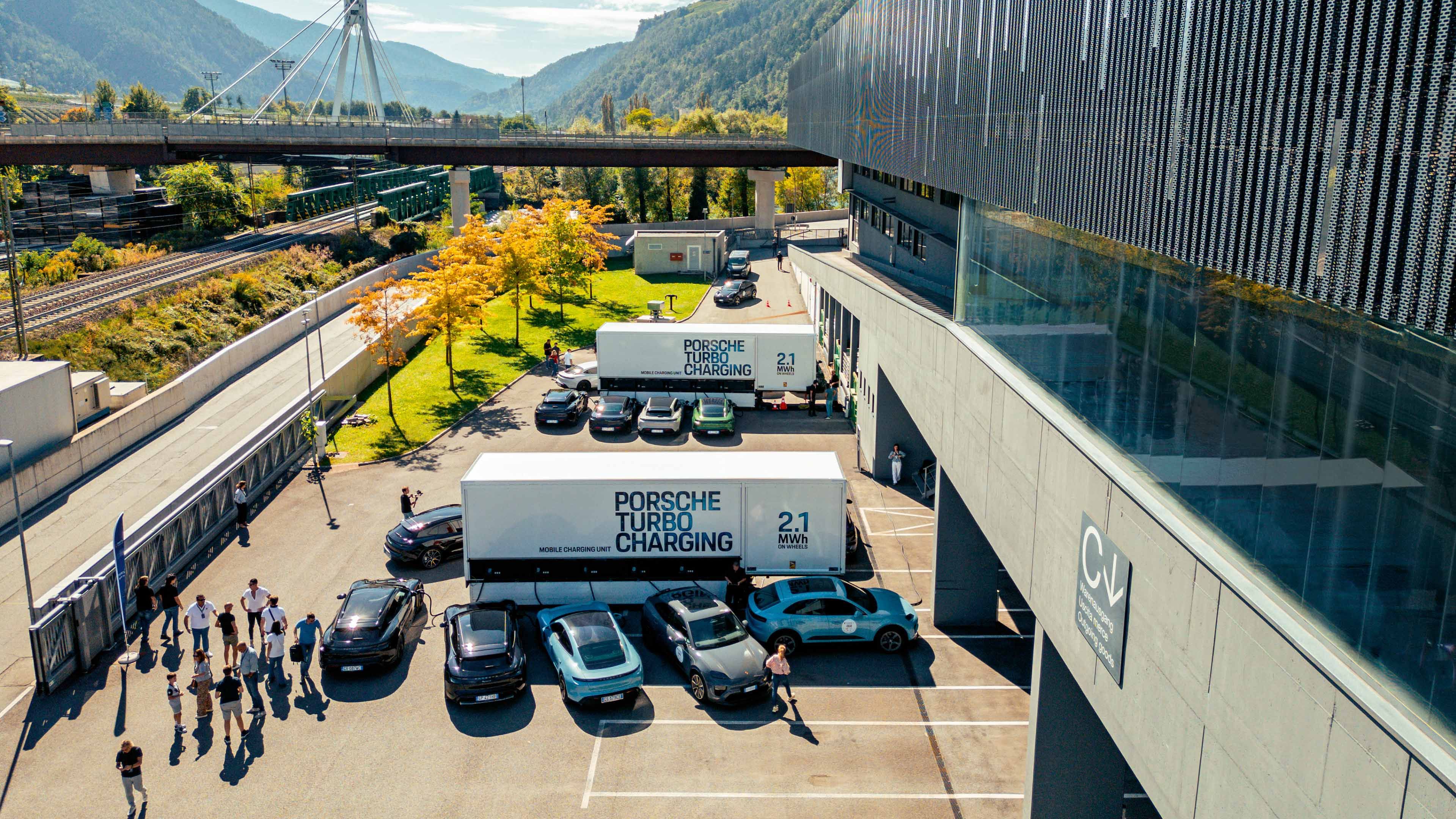 Several electric Porsche vehicles are parked in front of a modern building while people walk between two mobile “Porsche Turbo Charging” units.