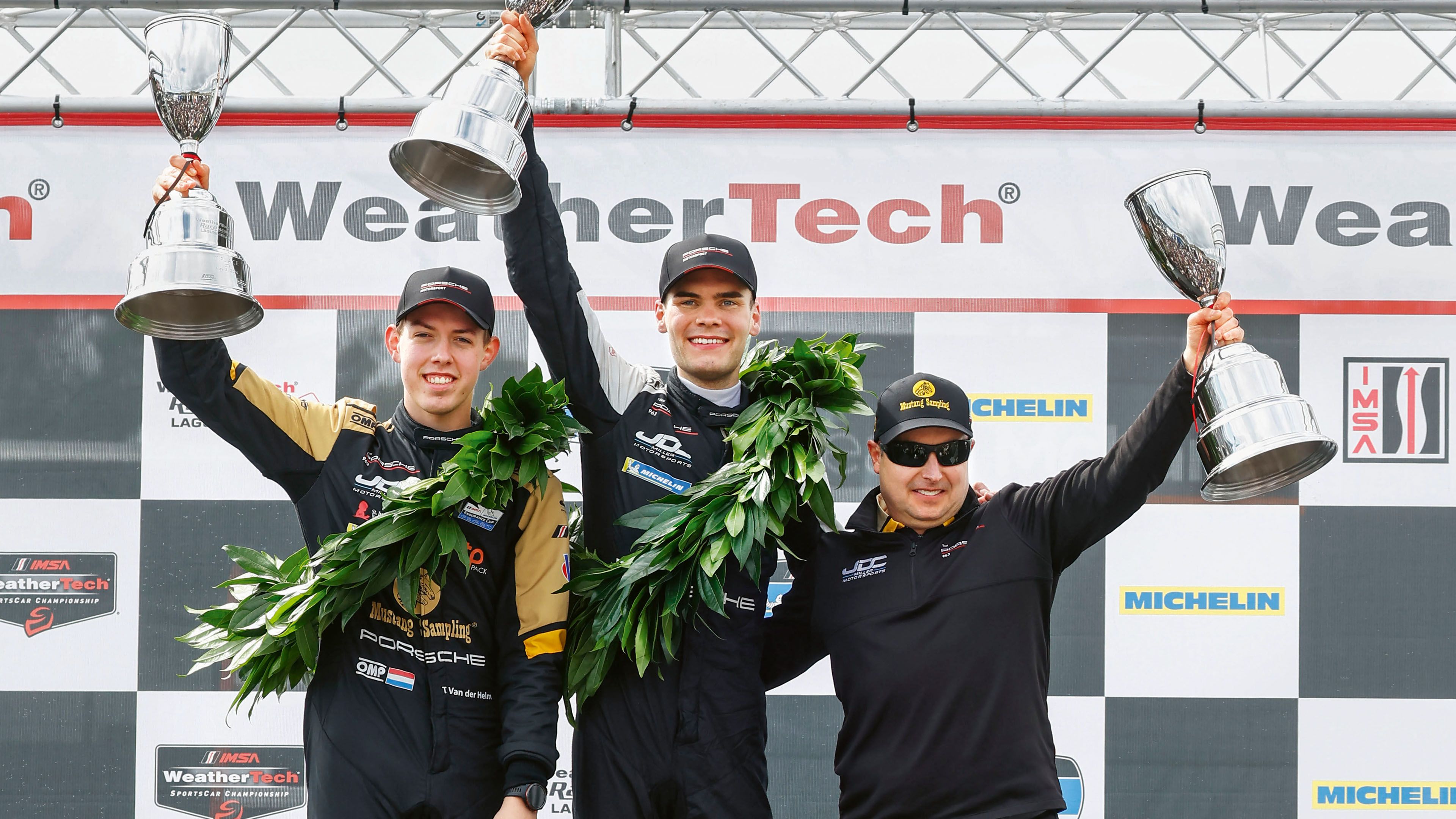 Three members of a racing team are standing on the podium at the podium ceremony for the IMSA race at Laguna Seca, holding trophies aloft.