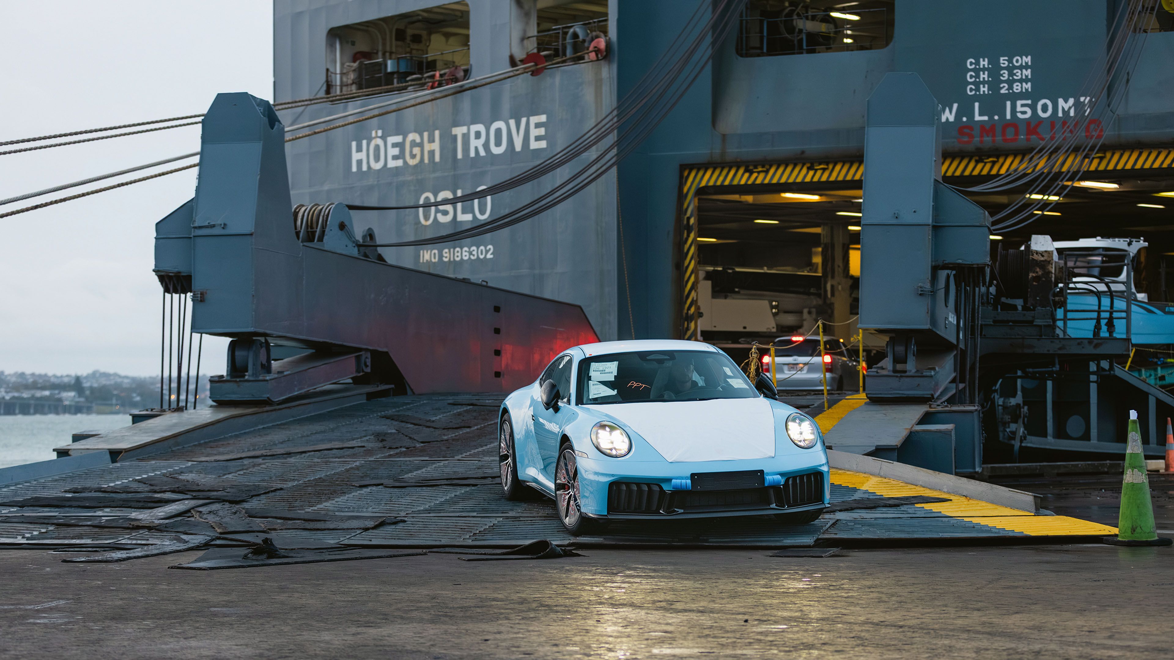 A Porsche 911 driving out of the cargo hold of a vehicle transport ship onto a harbour ramp.