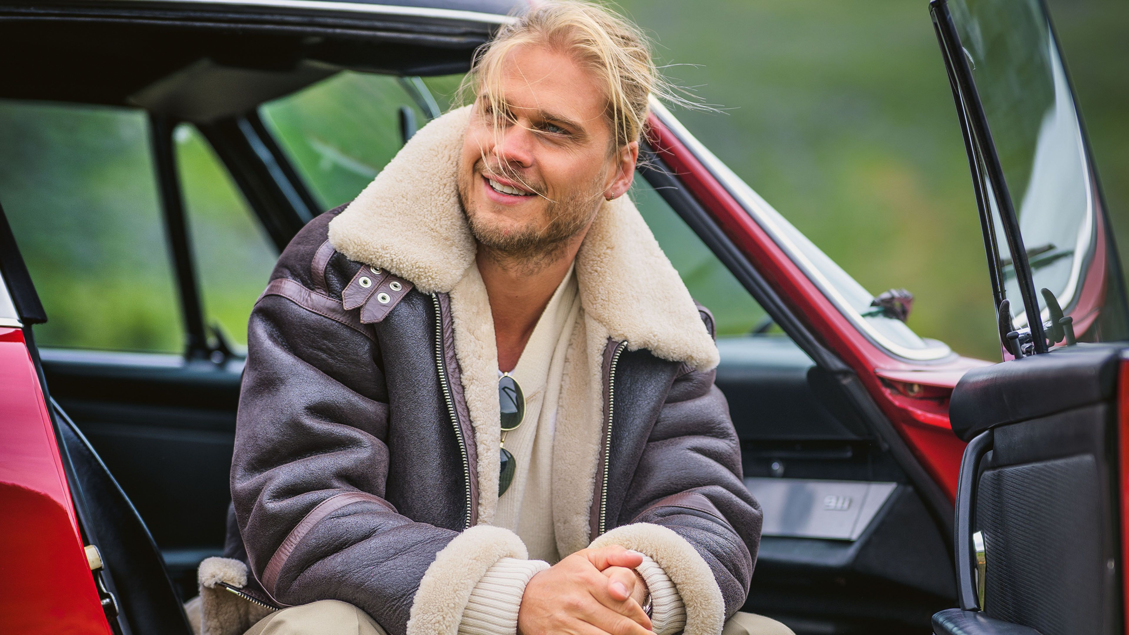 A young man with blond hair and a brown leather jacket sits smiling in the open doorway of a red 911 classic car.
