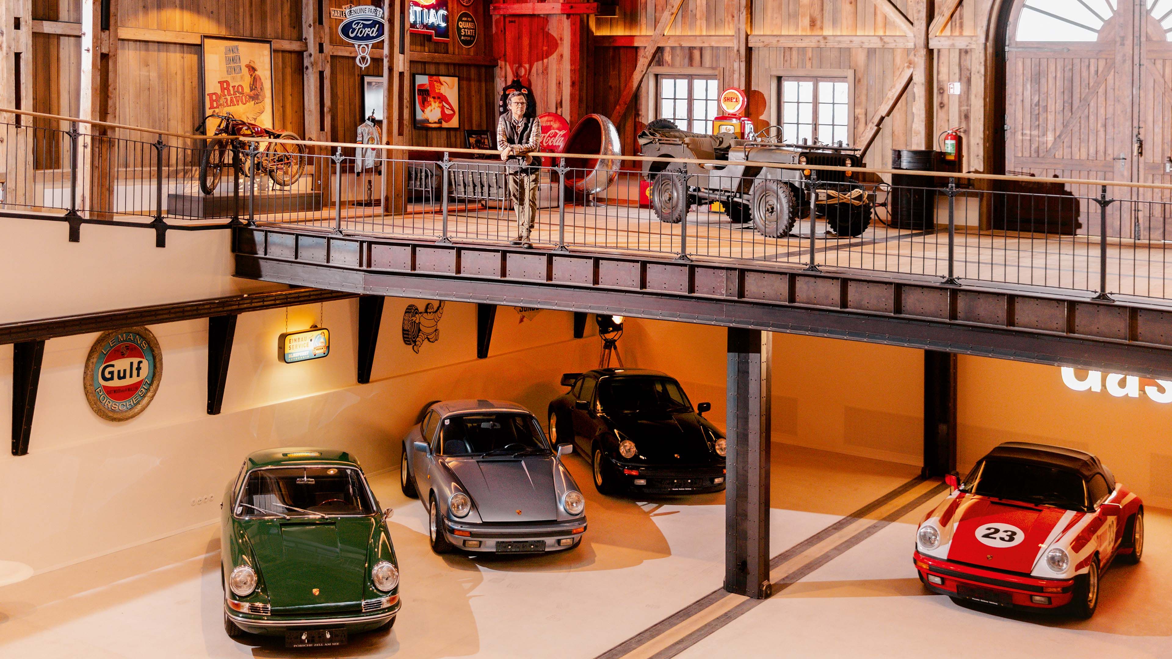 A vintage-themed garage housing a collection of Porsche 911 classic cars, with a person standing on the upper balcony, with various Porsche models displayed.