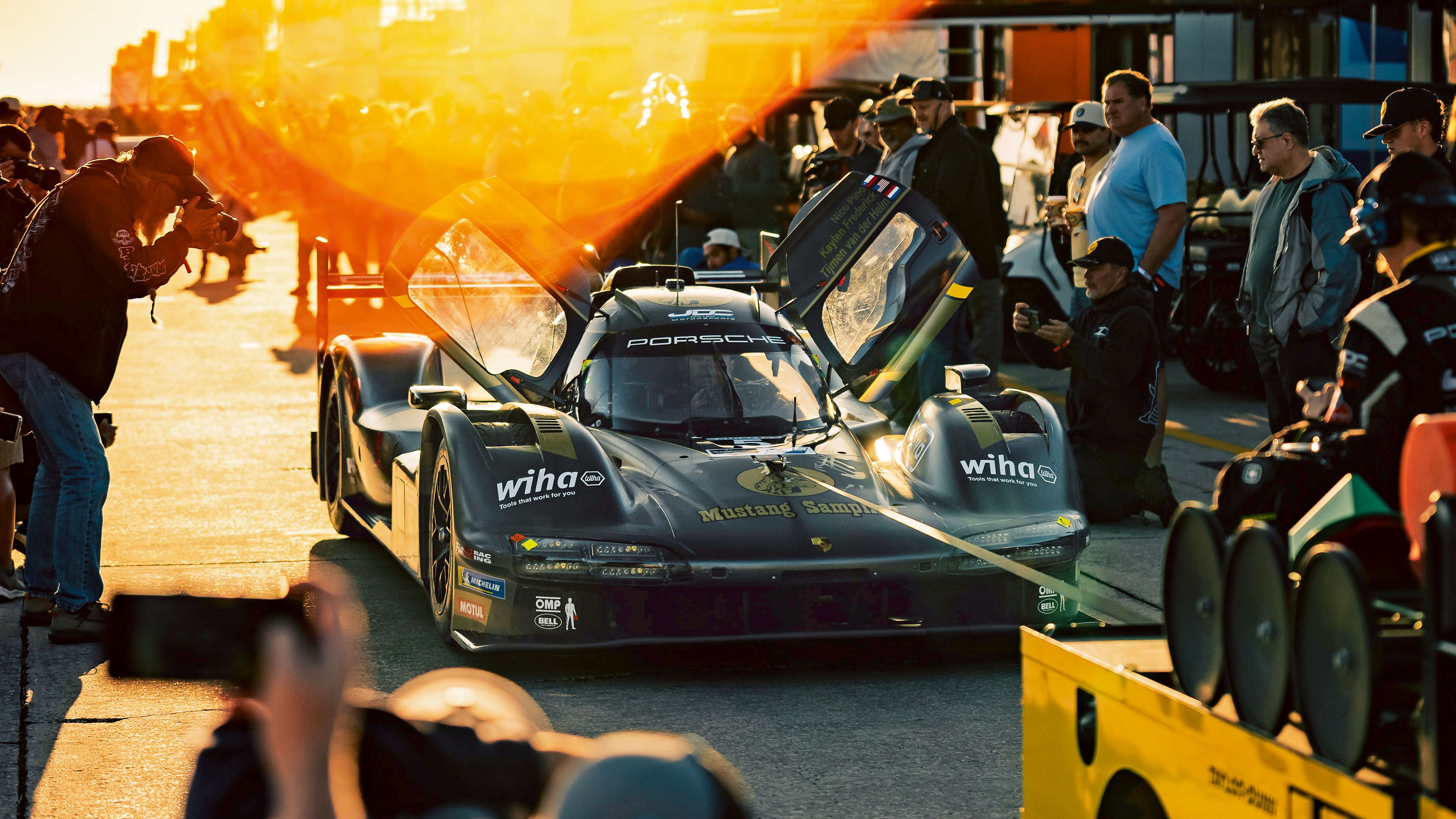 A Porsche race car with open doors is surrounded by team members, photographers, and spectators in the pit lane during sunset.