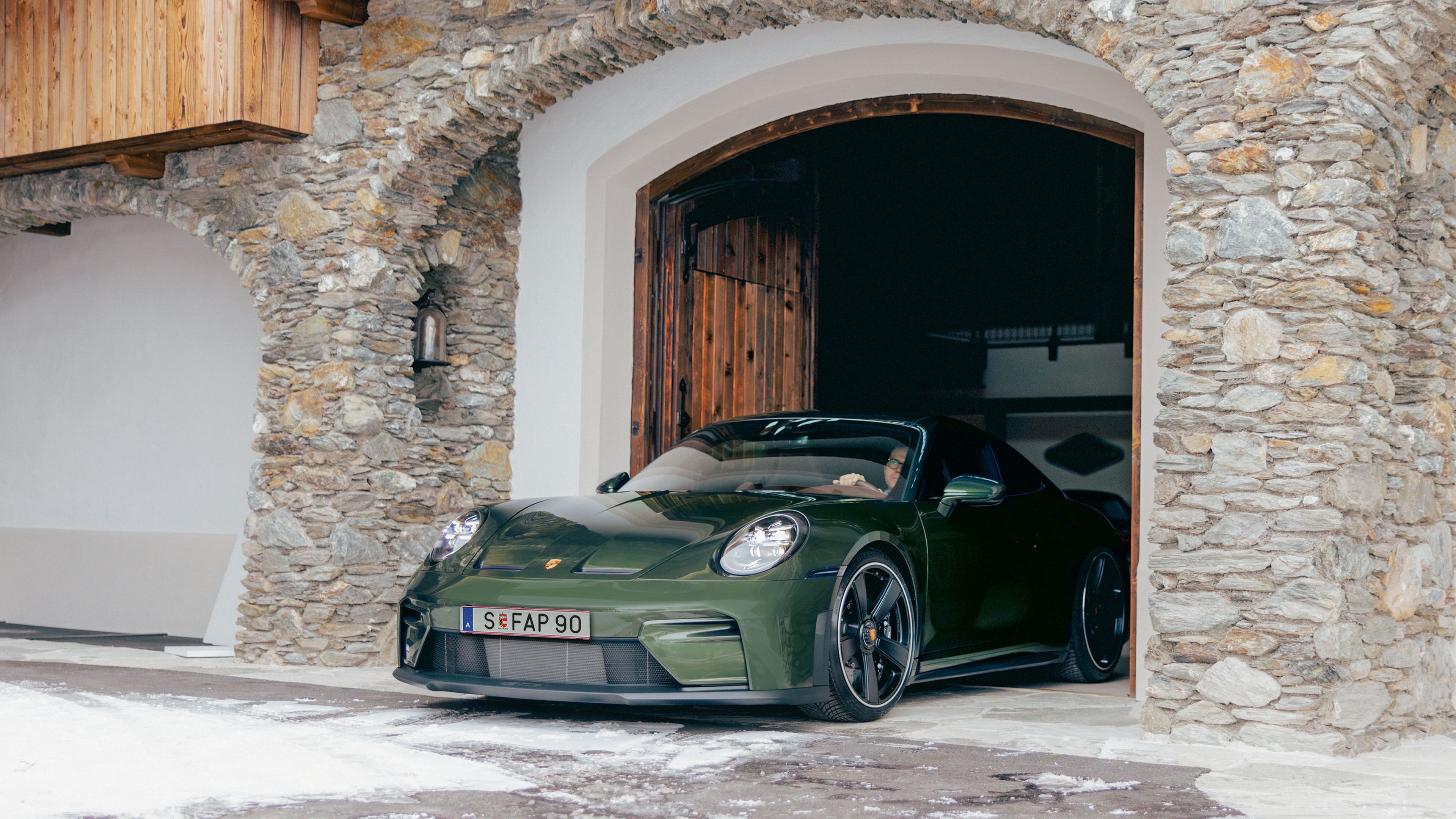 A dark green Porsche 911 is parked in front of a stone garage entrance, with snow on the ground and wooden structures in the background.