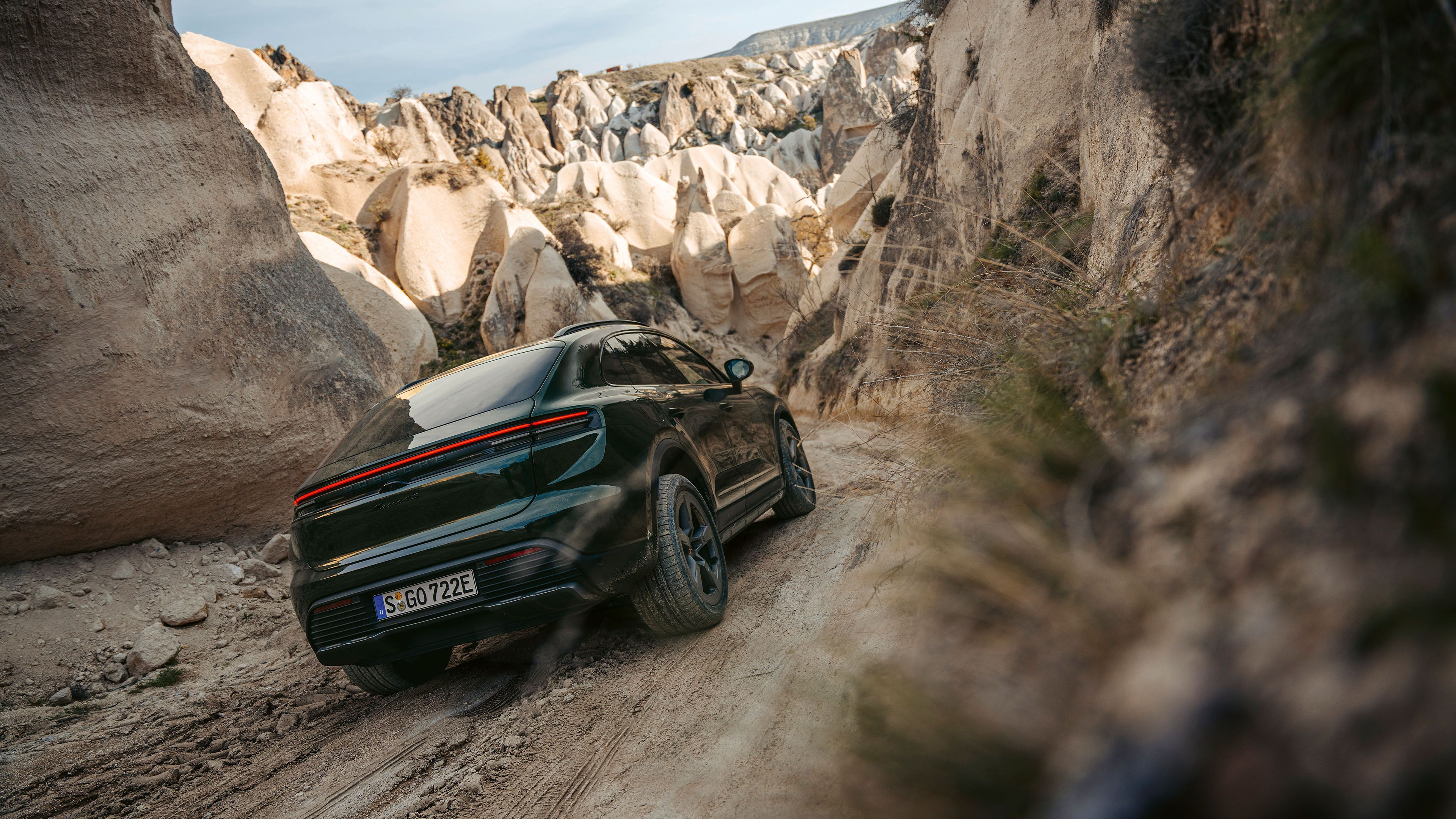 A dark green Porsche Macan 4S is driving along a narrow, rocky trail between high rock formations.