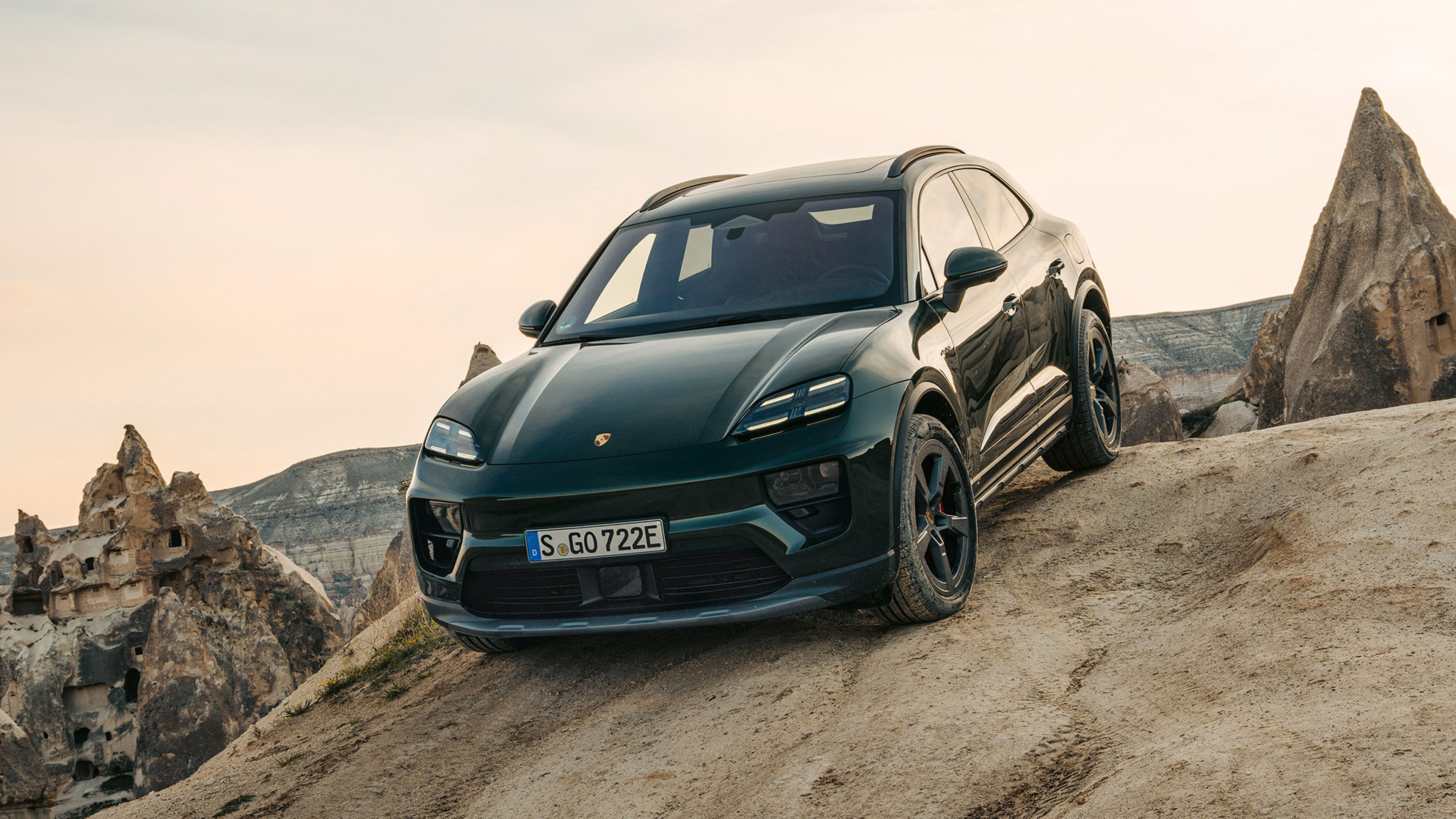 A dark green Porsche Macan 4S drives down a steep, sandy slope in front of a rocky, barren landscape under a cloudy sky.
