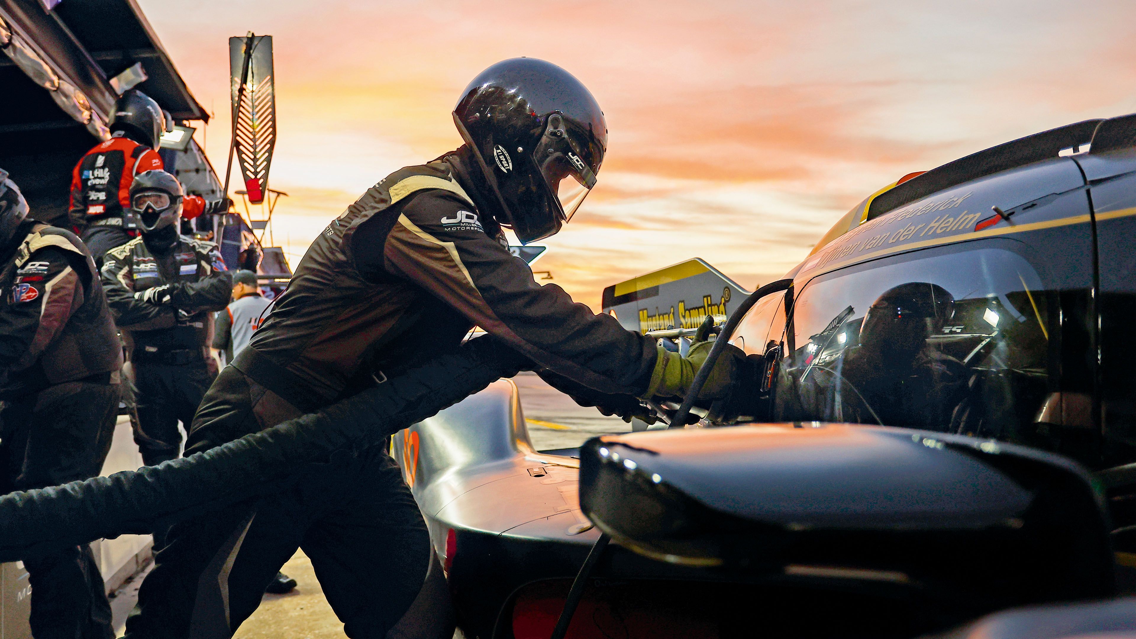 A pit crew member leans into a race car during a pit stop while other team members stand nearby at sunset.