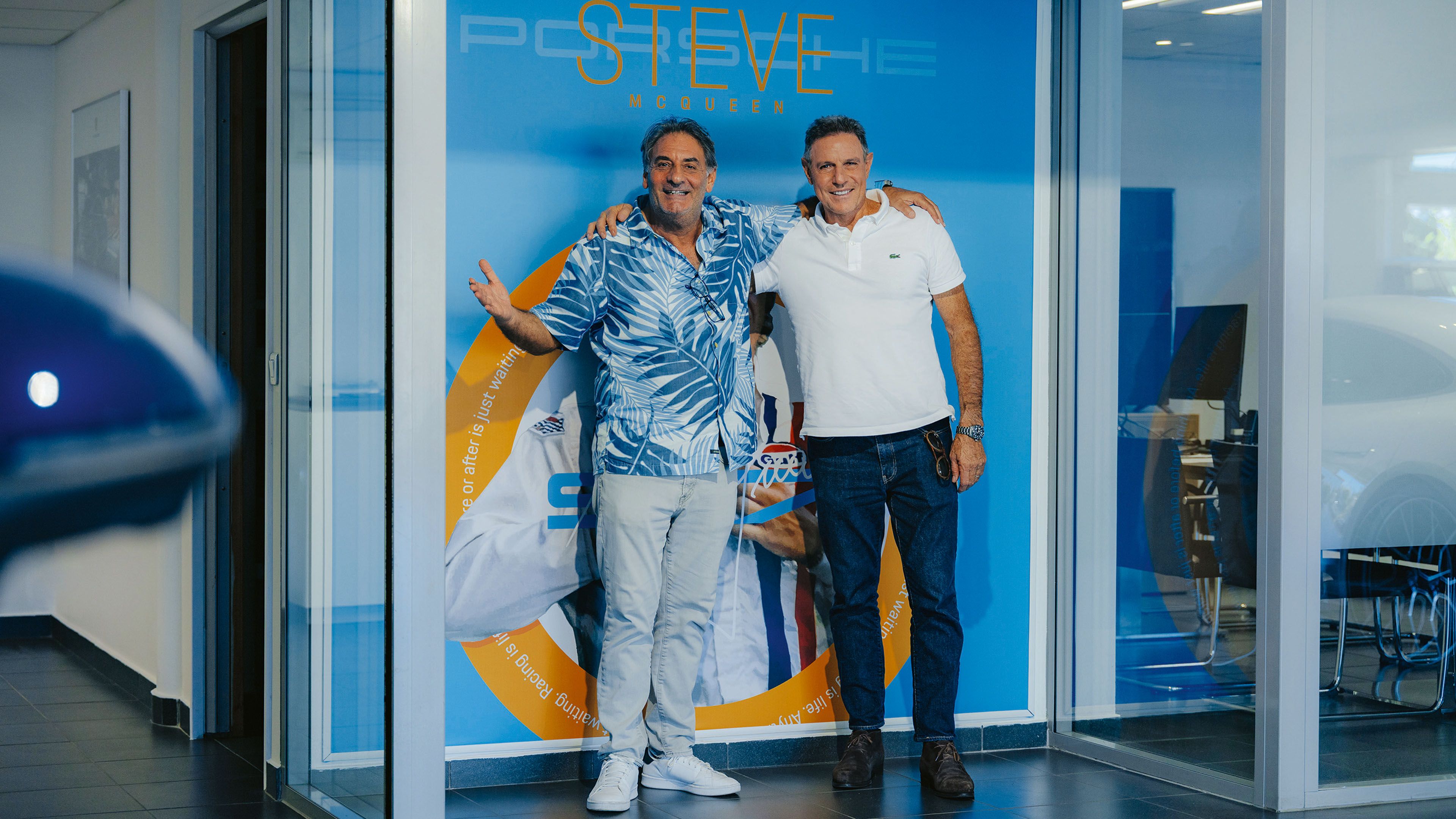 Two men standing in front of a Porsche exhibition area with blue wall design and a car in the background.
