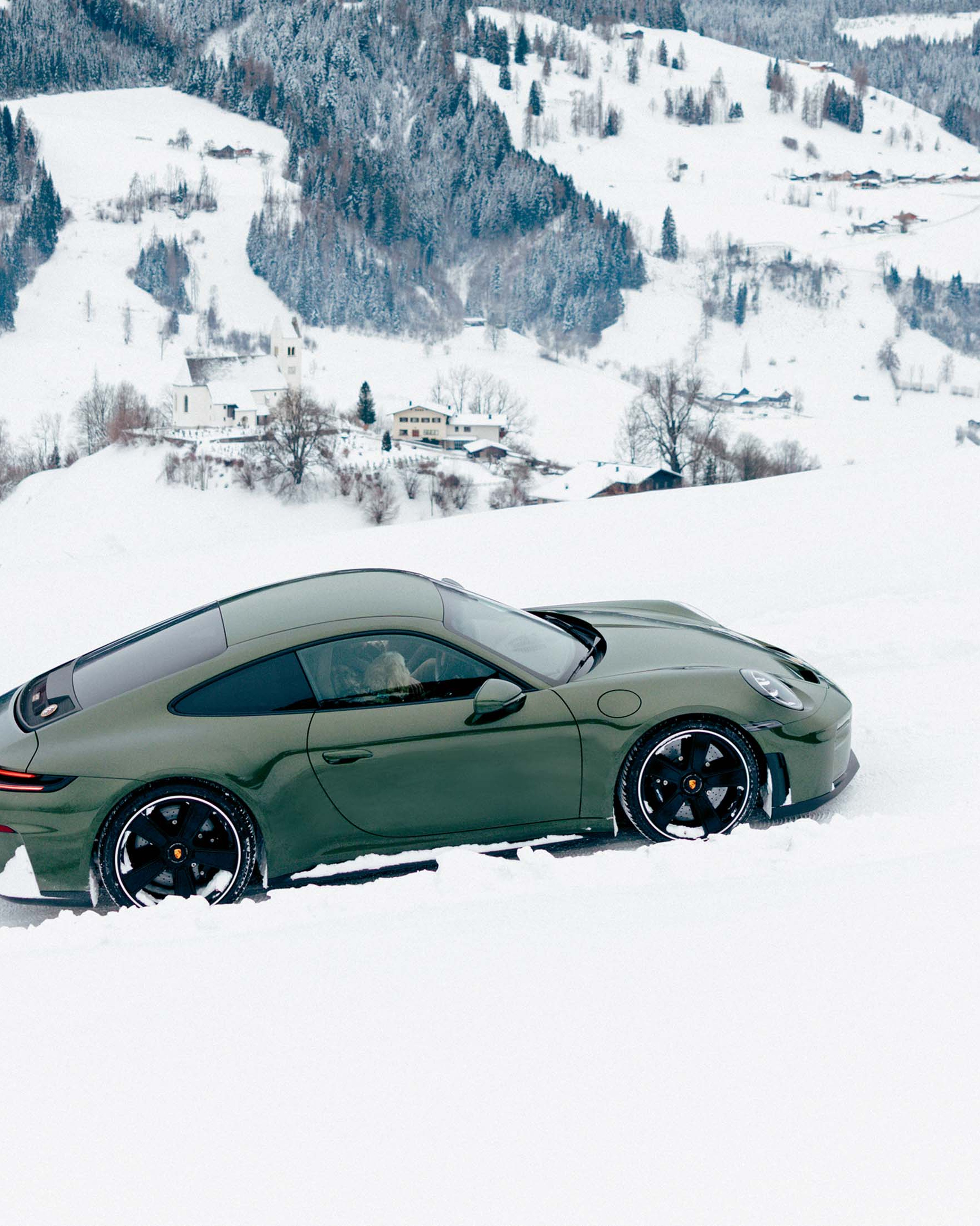 A dark green Porsche 911 drives through snowy terrain against the backdroph of a mountainous winter landscape.
