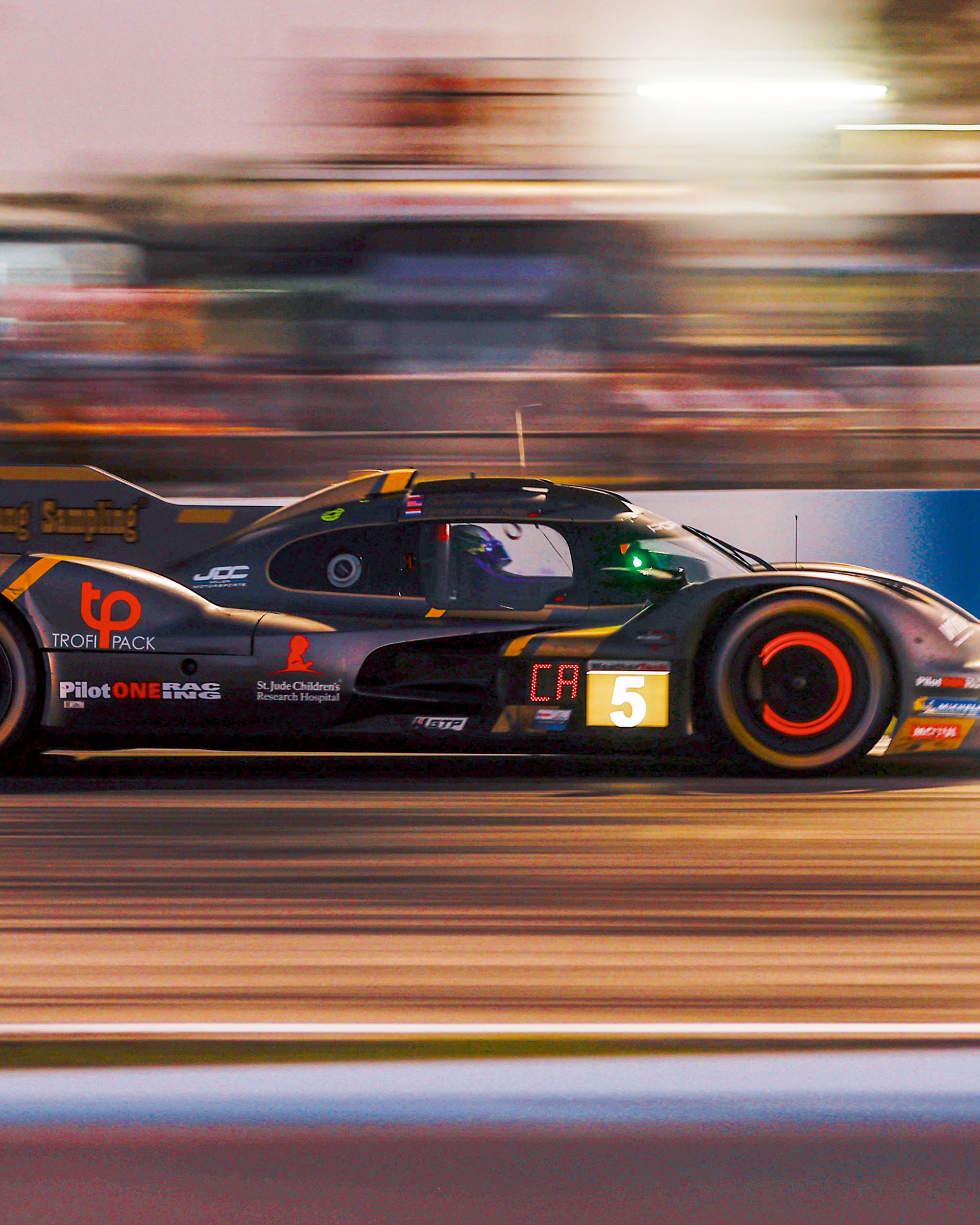 A Porsche race car drives along a racetrack at dusk with motion blur in the background.