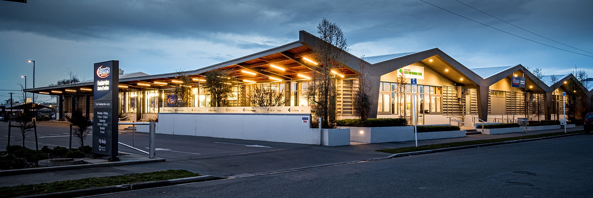Trents wholesale building, a modern low-slung commercial building with glowing warm interior lights and parking lot at dusk.