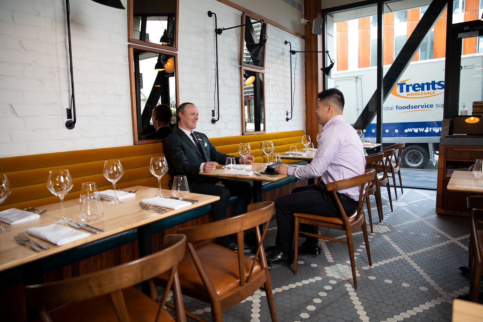 Two businessmen sit across a table in a modern restaurant, set wine glasses and large window.