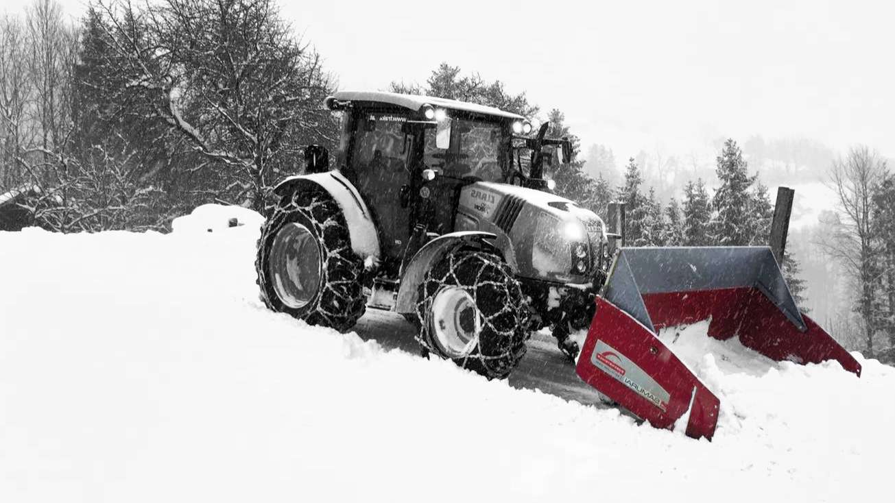A tractor with a snowplow attachment clears snow on a hilly, tree-lined terrain during heavy snowfall.