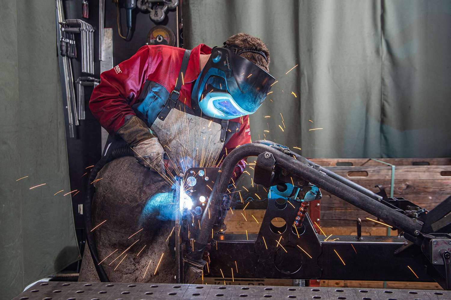 A welder in protective gear works, emitting bright sparks, in a workshop with metal frames and tools around.