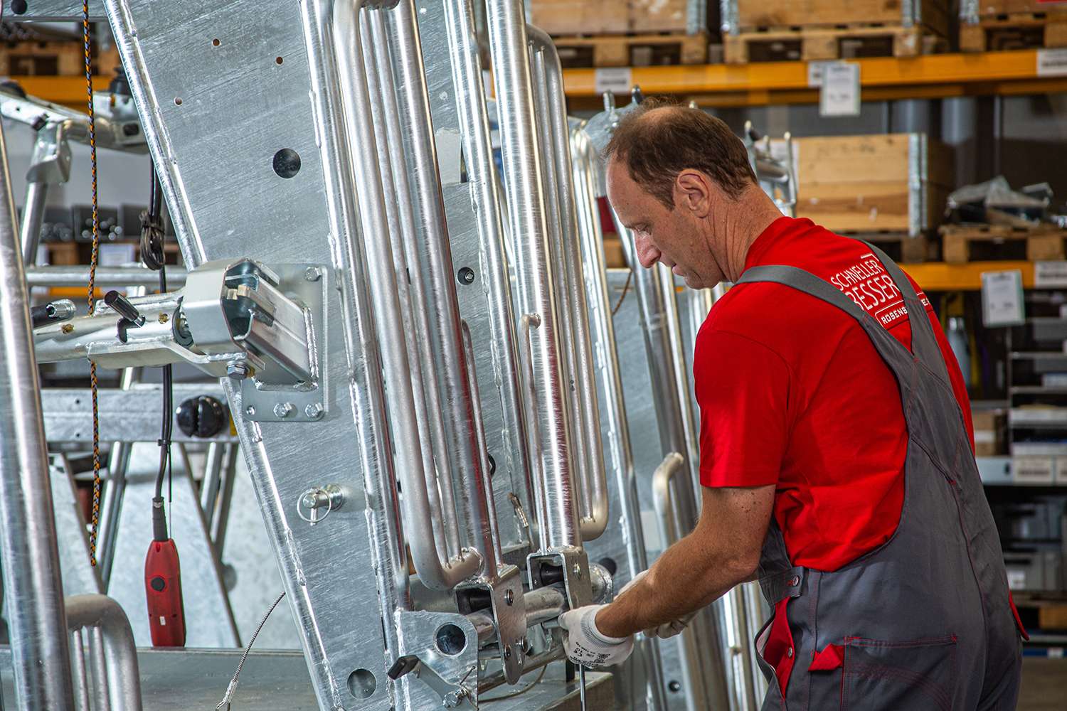 Man in red shirt and overalls working on industrial machinery in a warehouse. Shelves with wooden boxes are in the background.