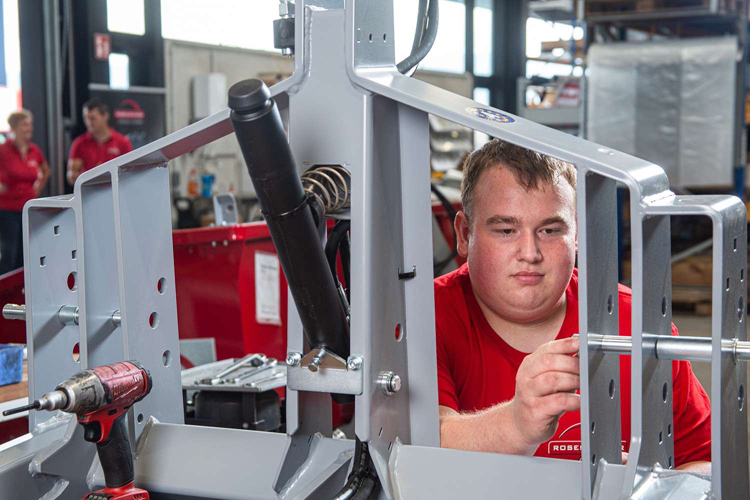 A person in a red shirt works on assembling machinery, surrounded by tools and workshop equipment.