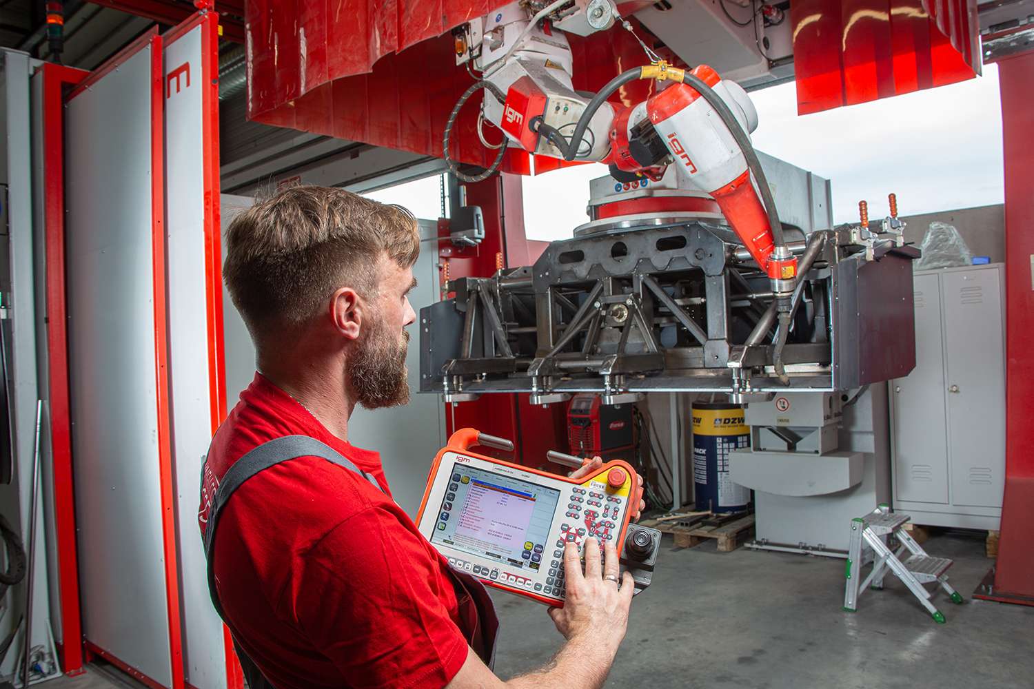A technician operates a robotic machine with a handheld control panel, inside an industrial setting with red equipment and metal structures.