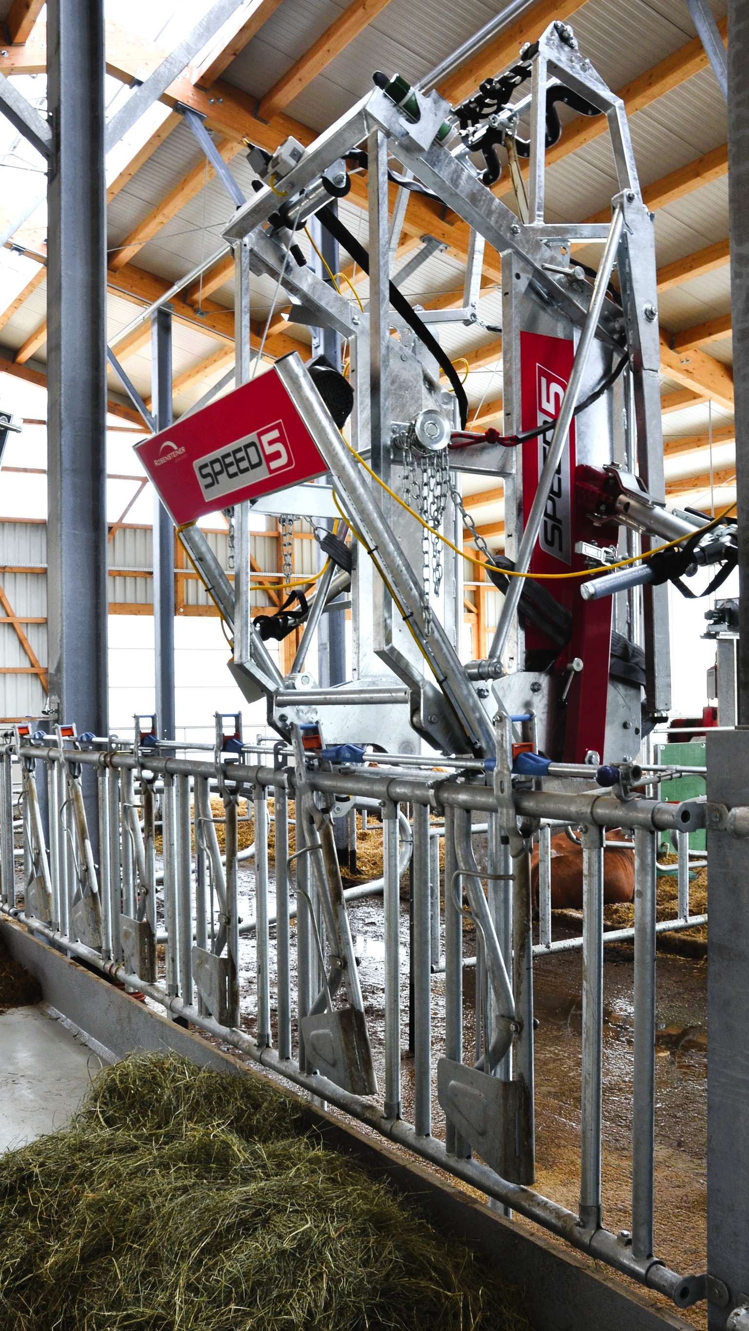 A metal livestock feeding machine in a barn, labeled "SPEED 5," positioned beside a feeding trough with hay.