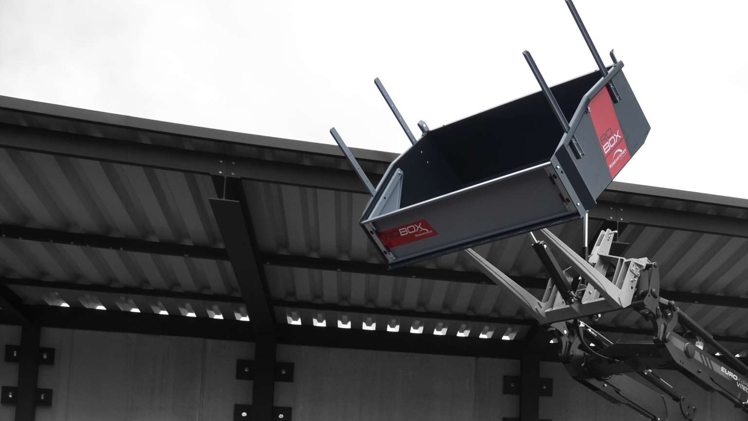 A large industrial machine lifting an angled metal container against a cloudy sky, with a corrugated metal roof in the background.