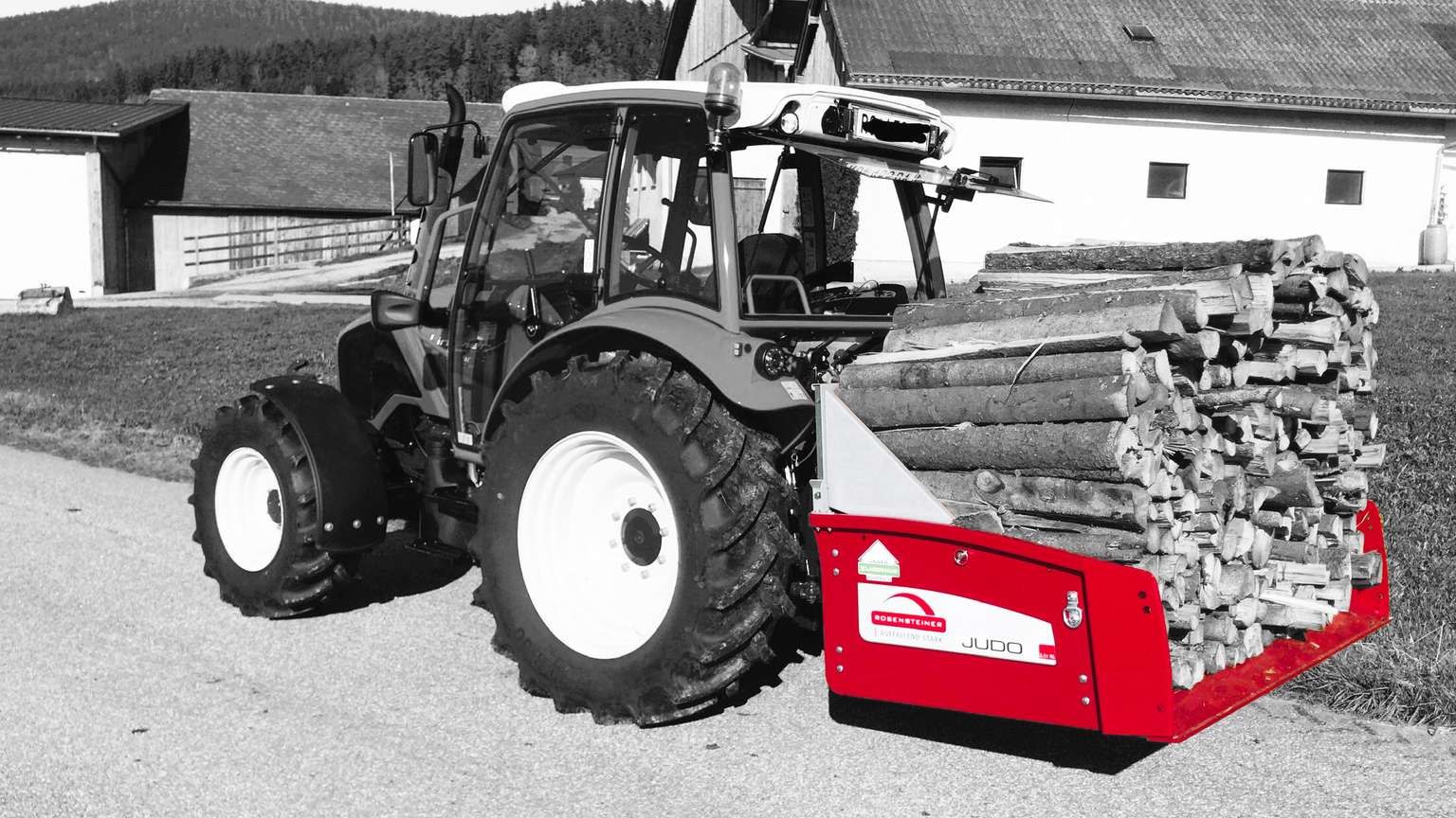 A tractor hauling a load of logs in a red attachment, parked on a rural road with farm buildings in the background.