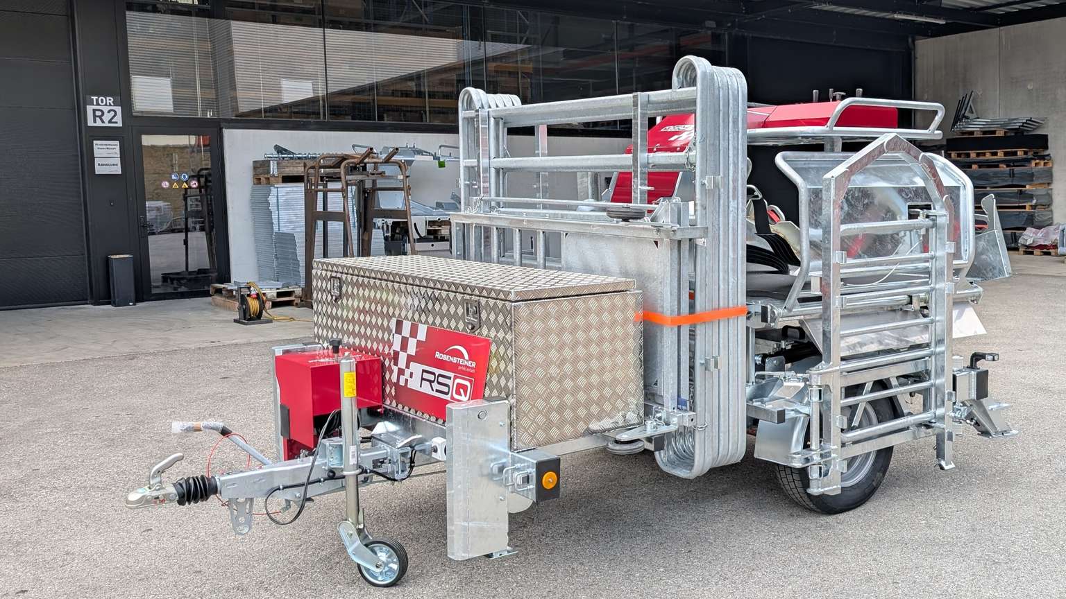 Industrial equipment trailer with metal framework and a red component, parked outside a warehouse with additional machinery nearby.