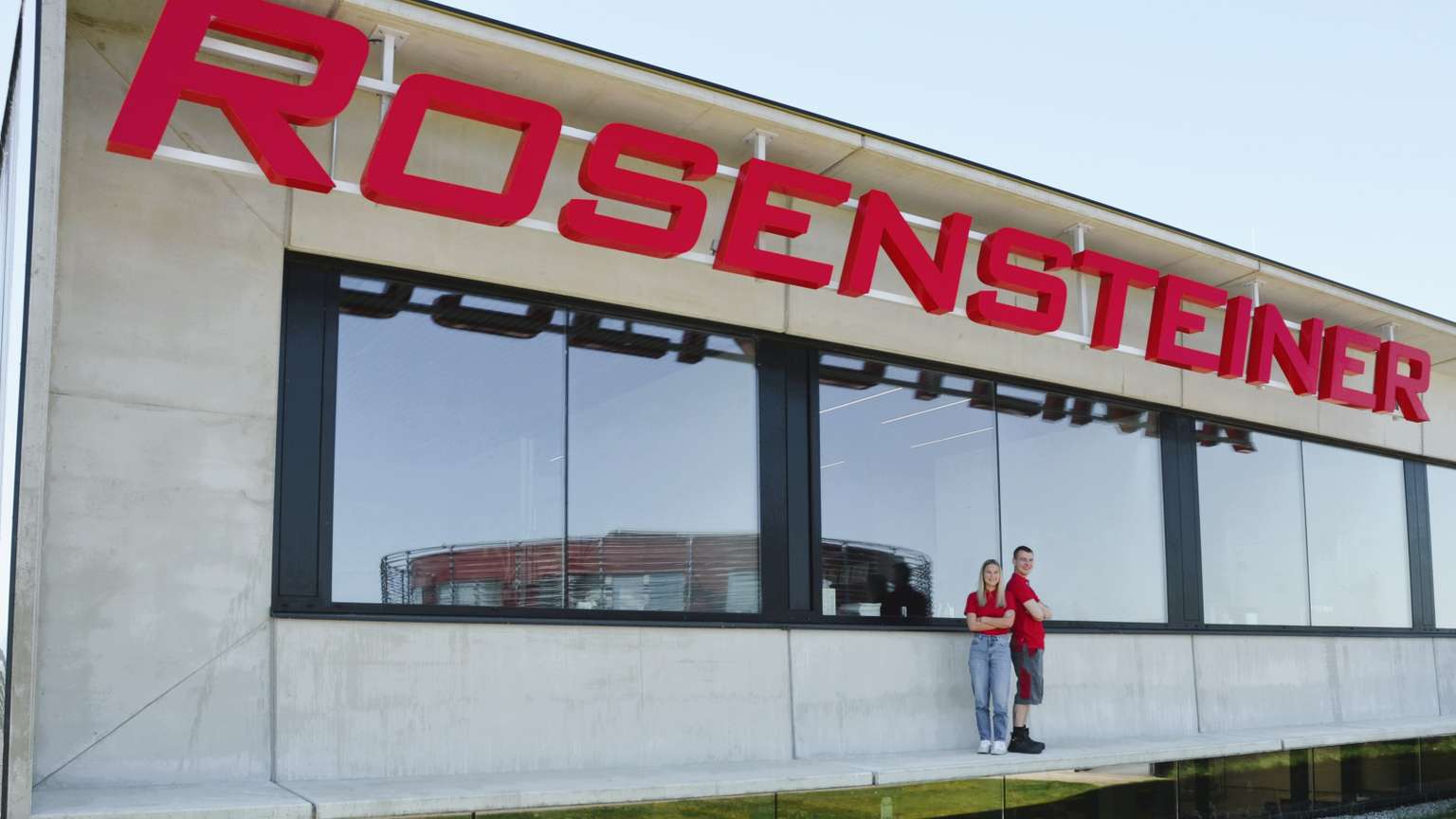 A large sign reading "ROSENSTEINER" on a modern building, with two people standing on a balcony, against a clear blue sky.